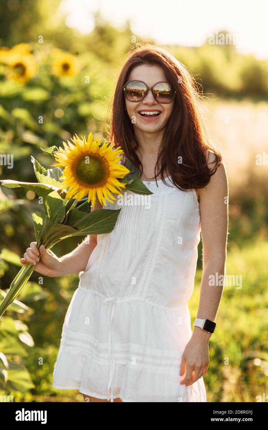 Bella giovane donna in un vestito bianco e in occhiali da sole in un campo con girasoli. Ritratto di donna in campagna al tramonto in estate giorno di sole Foto Stock