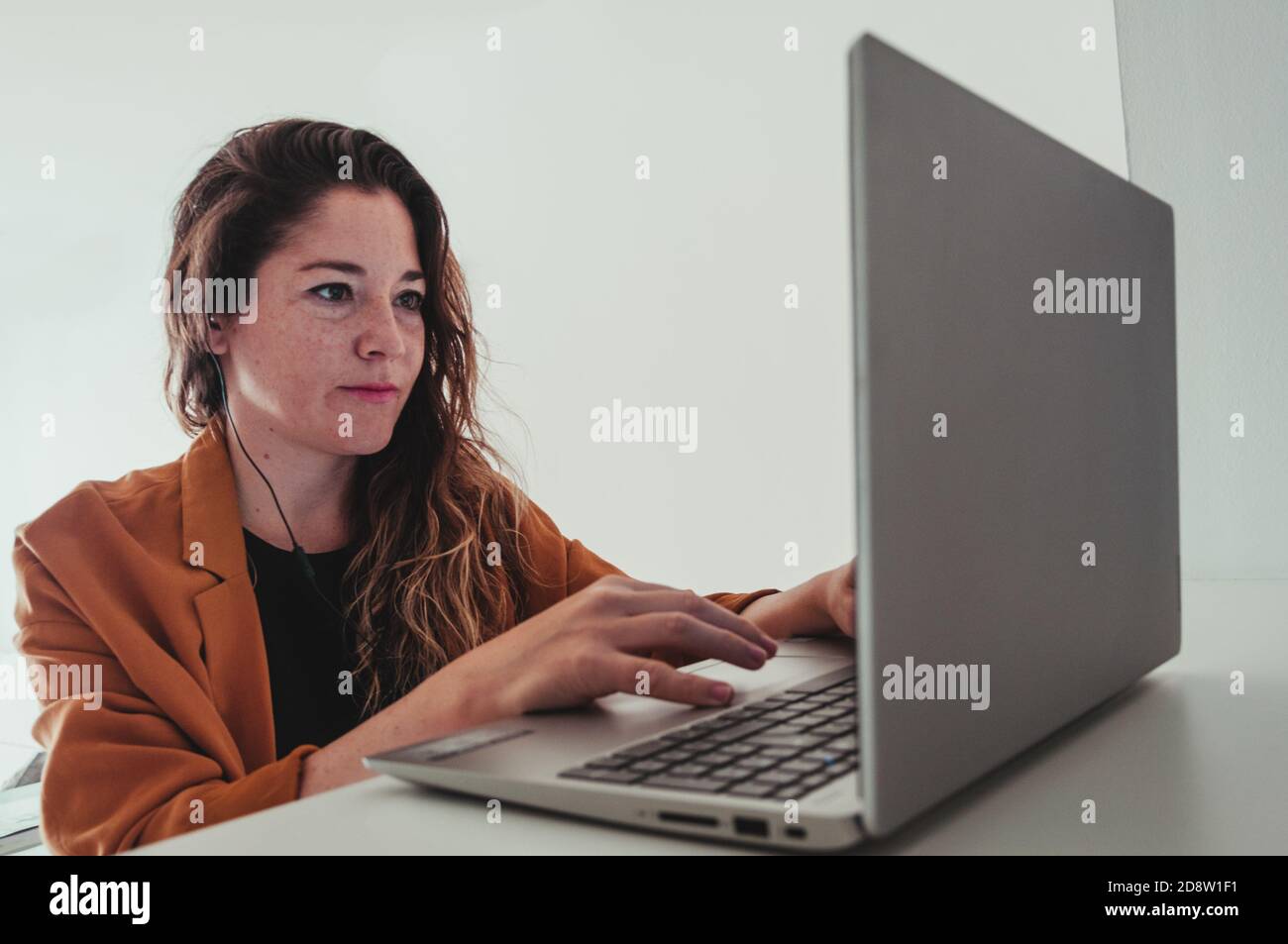 Bella donna con un computer portatile che lavora da casa. È seduta su una sedia in cucina a pianta aperta. Sta indossando le cuffie. Foto Stock