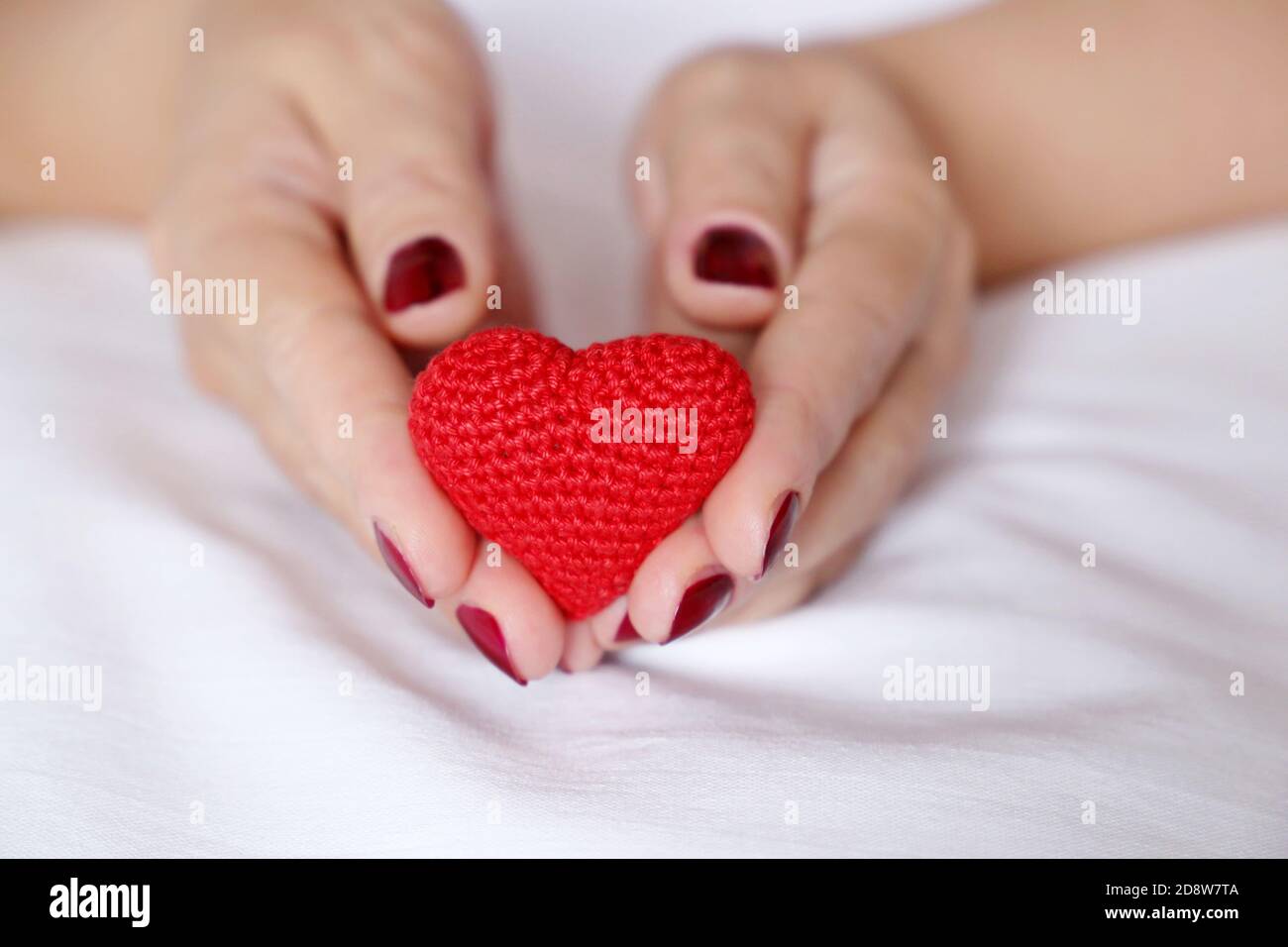 Cuore lavorato a maglia rosso in mani femminili su un letto. Concetto di amore, celebrazione dei San Valentino, maternità, donazione di sangue Foto Stock