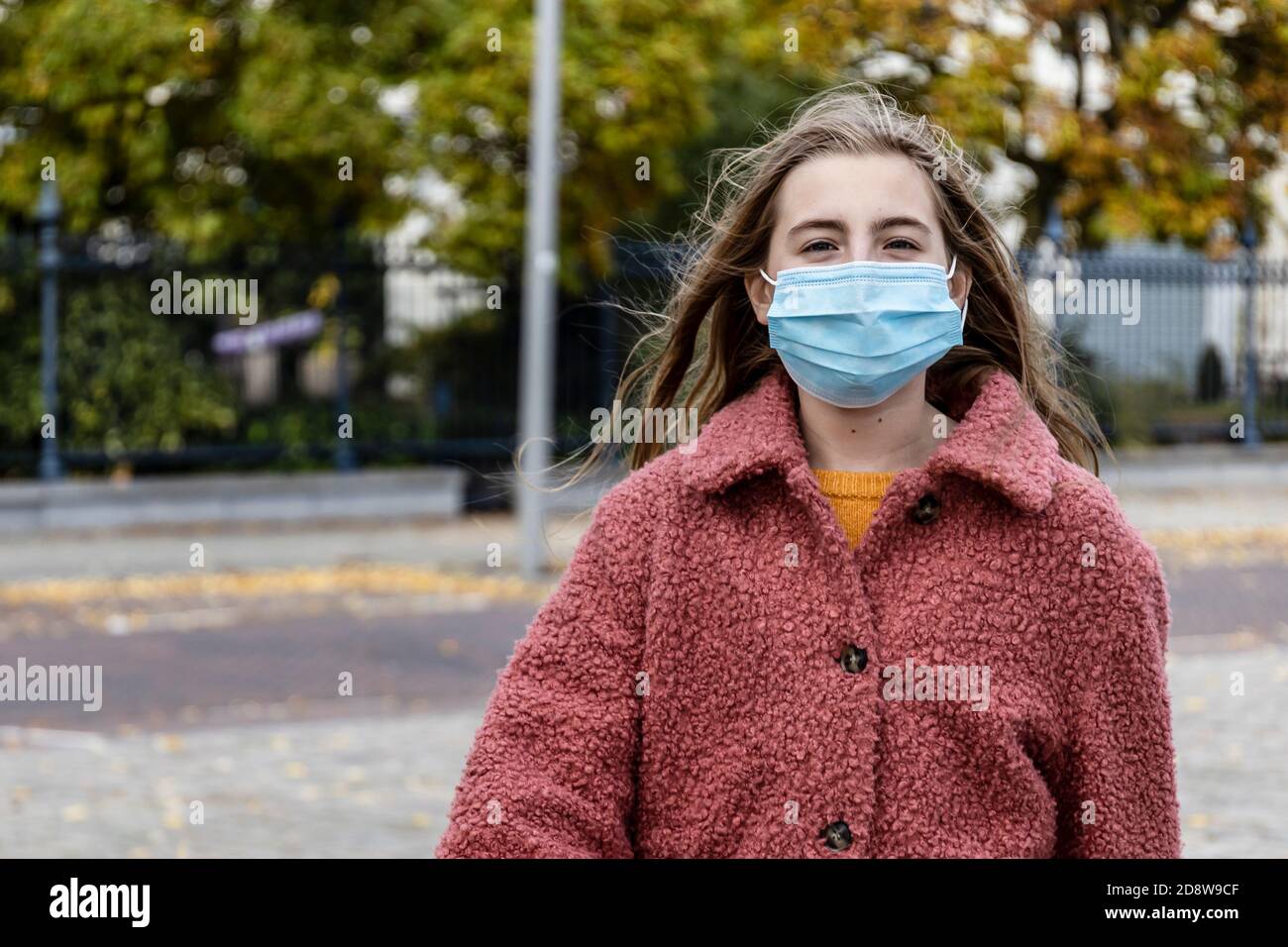 Bionda ragazza in camice rosa invernale camminare con fiducia su una strada della città indossare una maschera viso per la pandemia di Corona virus. Foto Stock