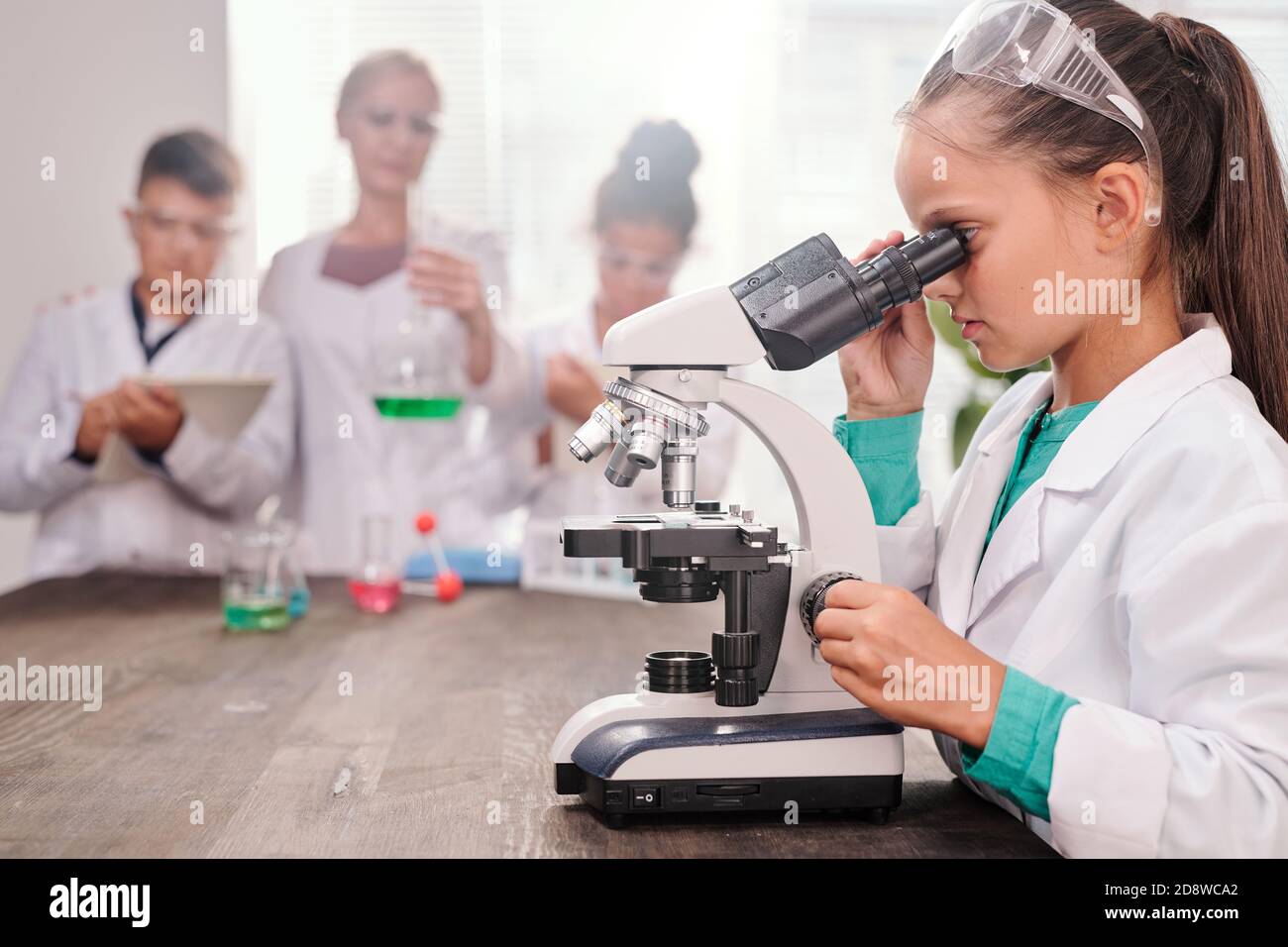 Curiosa studentessa in whitecoat guardando al microscopio da scrivania contro compagni di classe Foto Stock