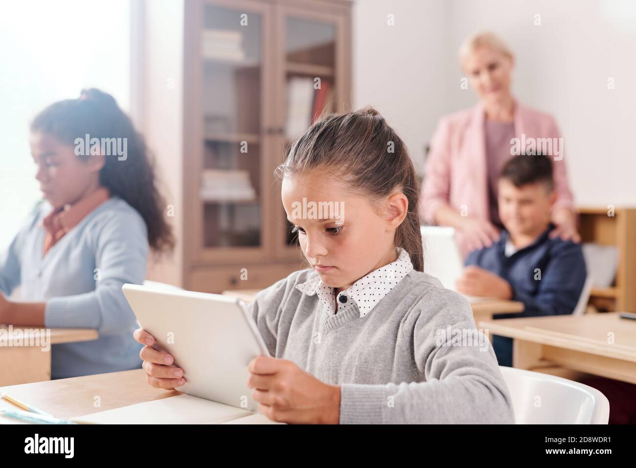 Carina ragazza e i suoi compagni di classe con tablet che lavorano individualmente con testo online Foto Stock