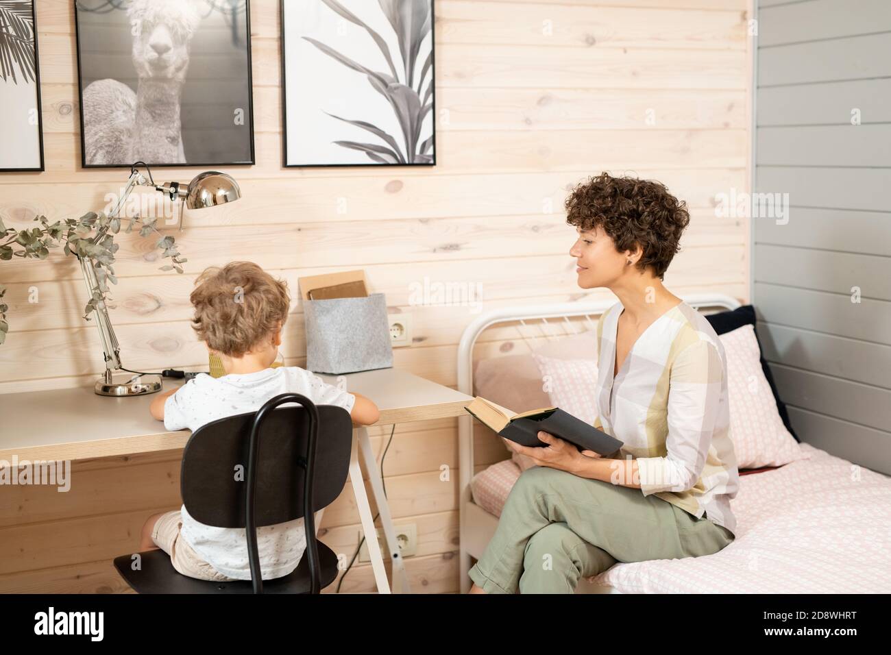 Giovane donna di brunette casual con libro aperto seduto sul letto da suo figlio piccolo Foto Stock
