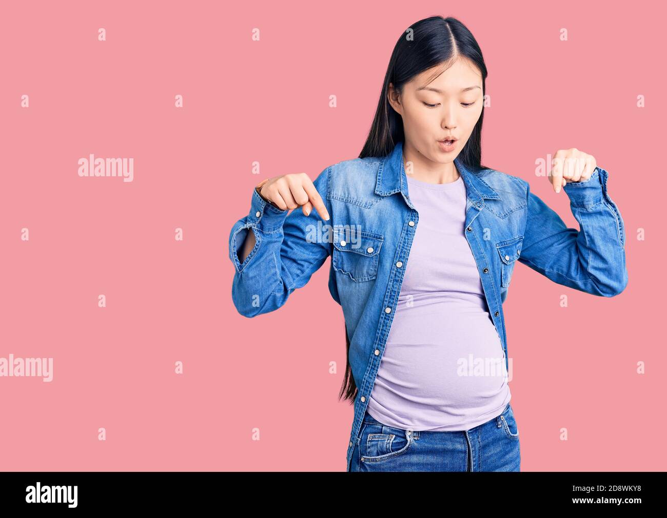 Giovane bella donna cinese incinta in attesa di bambino puntando con le dita che mostrano pubblicità, viso sorpreso e bocca aperta Foto Stock