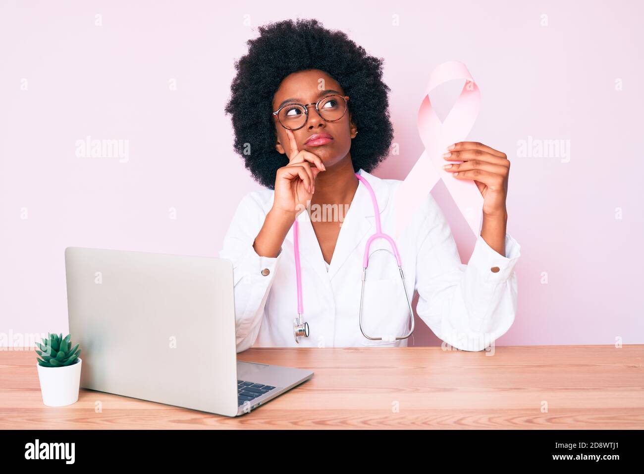 Giovane donna afroamericana che indossa uniforme medico che tiene il nastro di cancro rosa faccia seria che pensa alla domanda con la mano sul mento, pensieroso abou Foto Stock