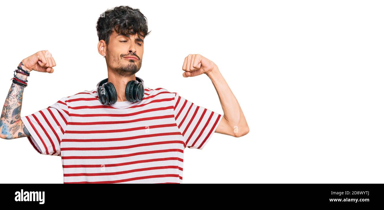 Giovane uomo ispanico che ascolta musica con le cuffie mostrando muscoli delle braccia sorridendo orgoglioso. Concetto di fitness. Foto Stock