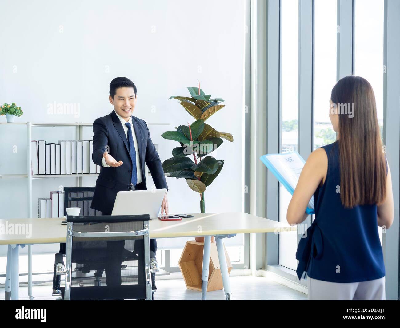 Uomo d'affari asiatico, capo nel vestito facendo gesto di mano per invitare la giovane donna che tiene il curriculum nell'intervista di lavoro a sedersi sulla sedia vicino vetro enorme Foto Stock