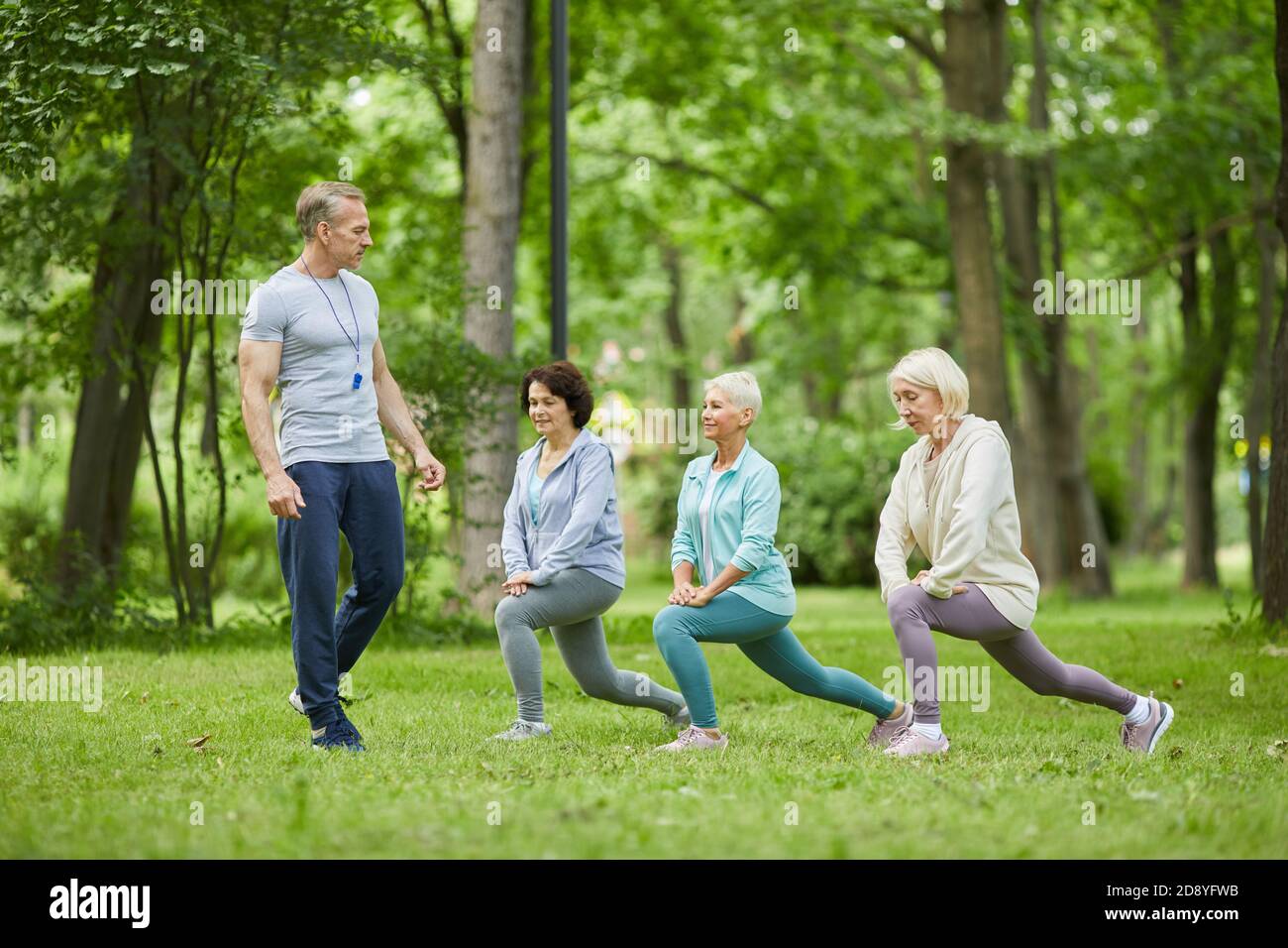 Ampio colpo di tre donne anziane buon-osservanti che hanno workout dentro parco cittadino con istruttore che li guarda Foto Stock