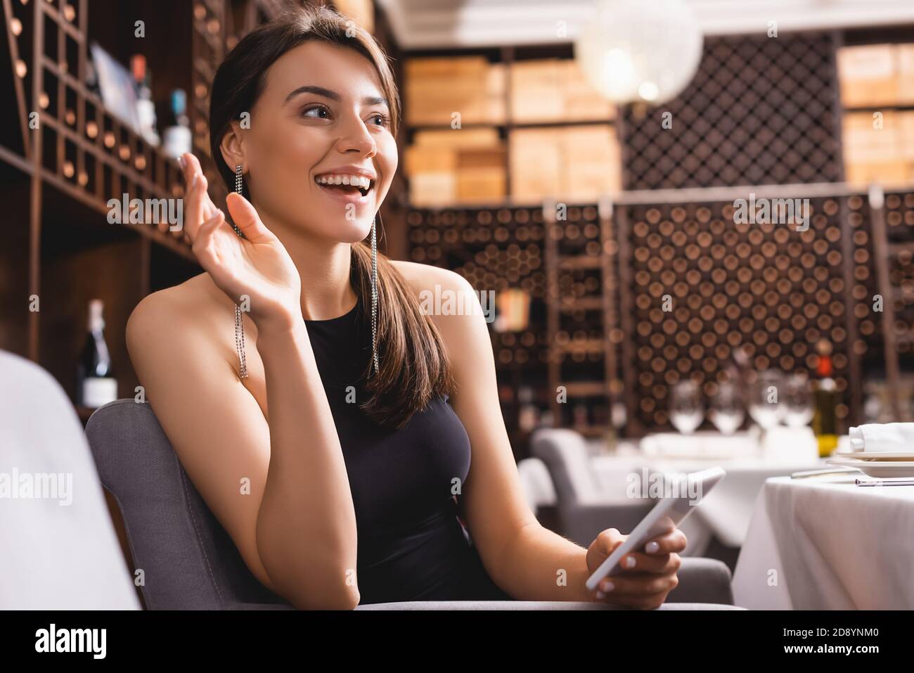 Messa a fuoco selettiva di una donna elegante che agita la mano mentre tiene lo smartphone nel ristorante Foto Stock