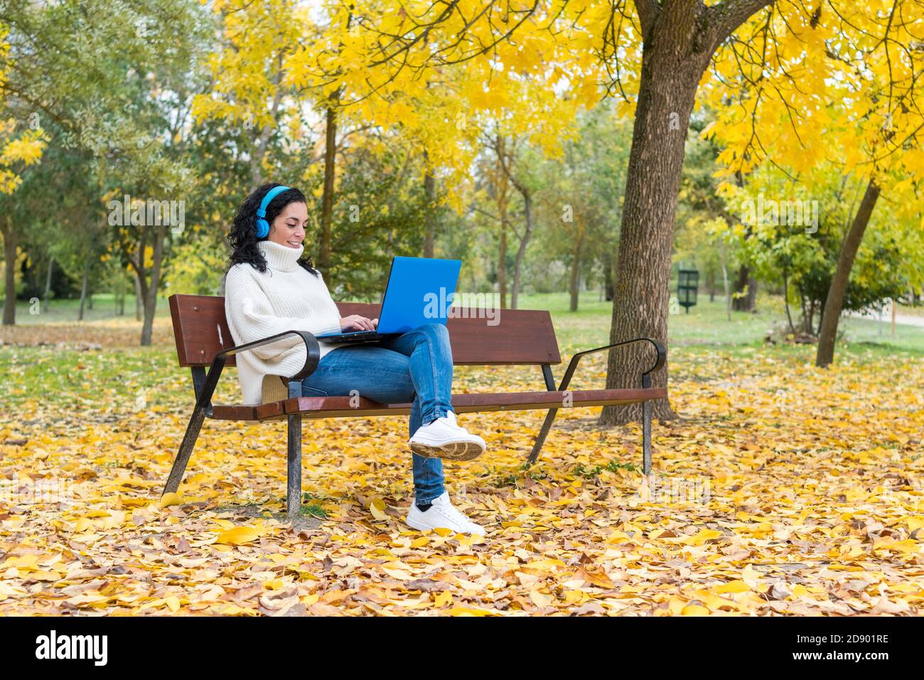 giovane donna con capelli ricci seduta su una panca di lavoro con computer portatile e cuffie wireless blu mentre sorride in autunno con foglie che cadono dal t Foto Stock