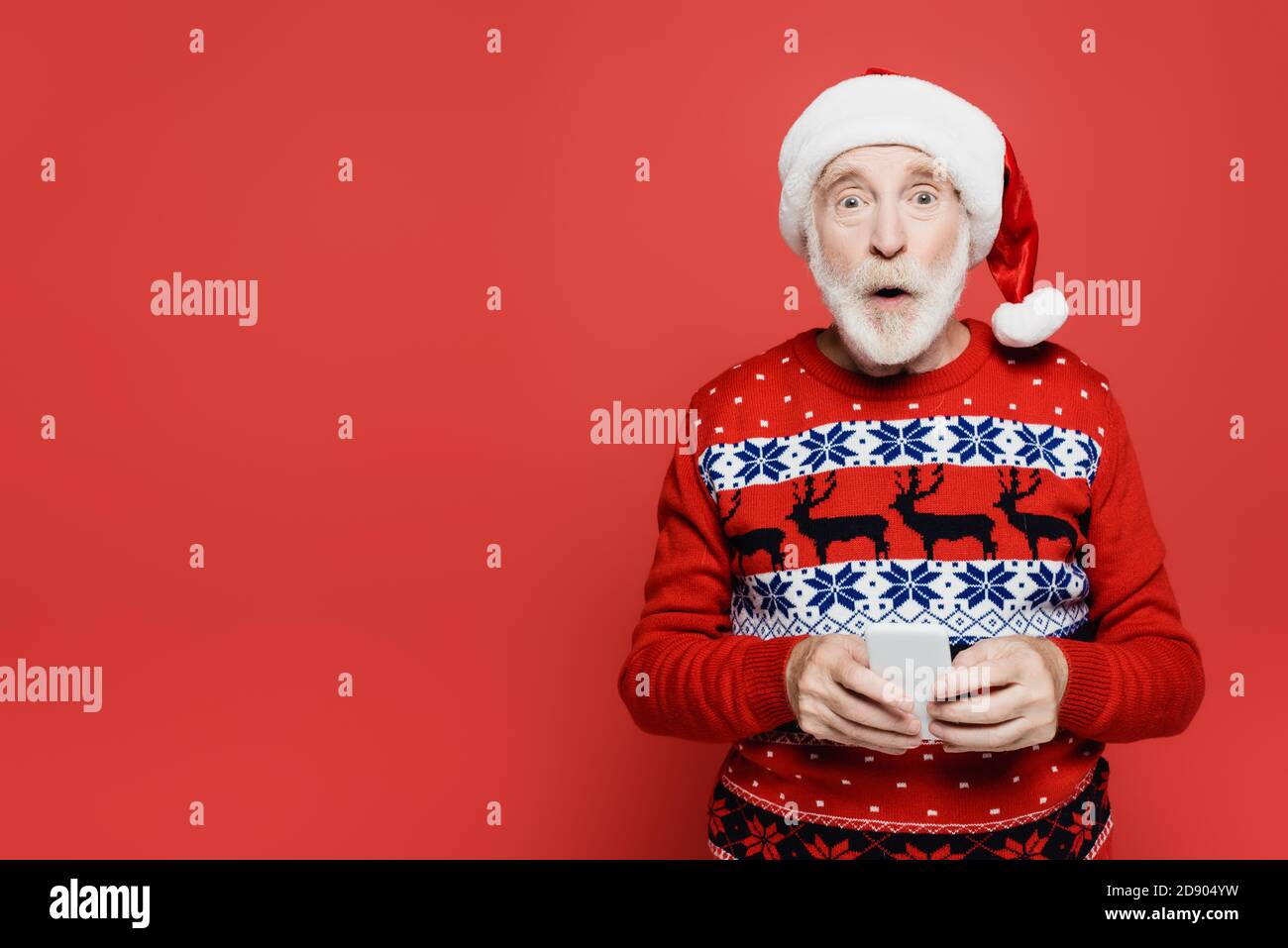 Uomo anziano scioccato in santa Hat che tiene lo smartphone isolato rosso Foto Stock