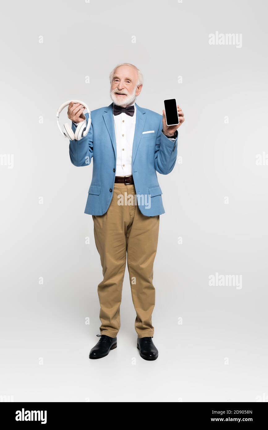 Uomo anziano sorridente che tiene le cuffie wireless e lo smartphone in grigio sfondo Foto Stock