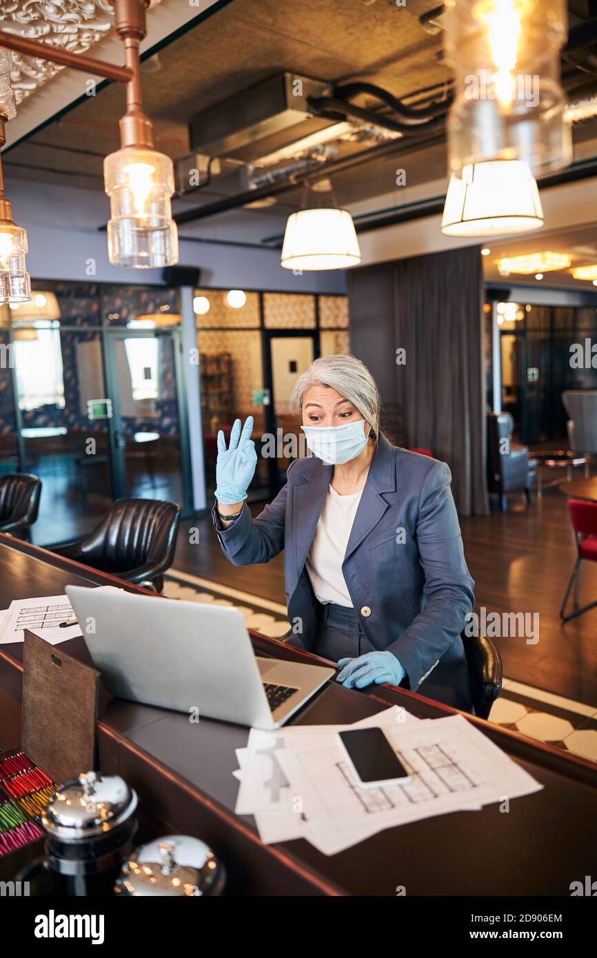 Uomo d'affari in maschera medica utilizzando notebook e facendo ciao gesto Foto Stock