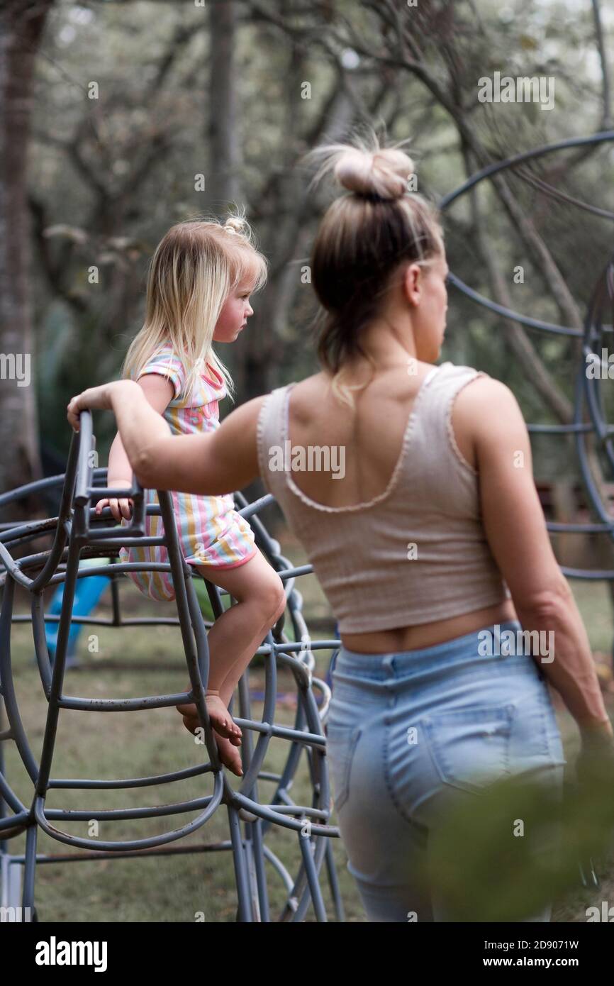 Una ragazza sale sulle barre della scimmia in un parco Come la sua nanny/Madre si trova nelle vicinanze Foto Stock
