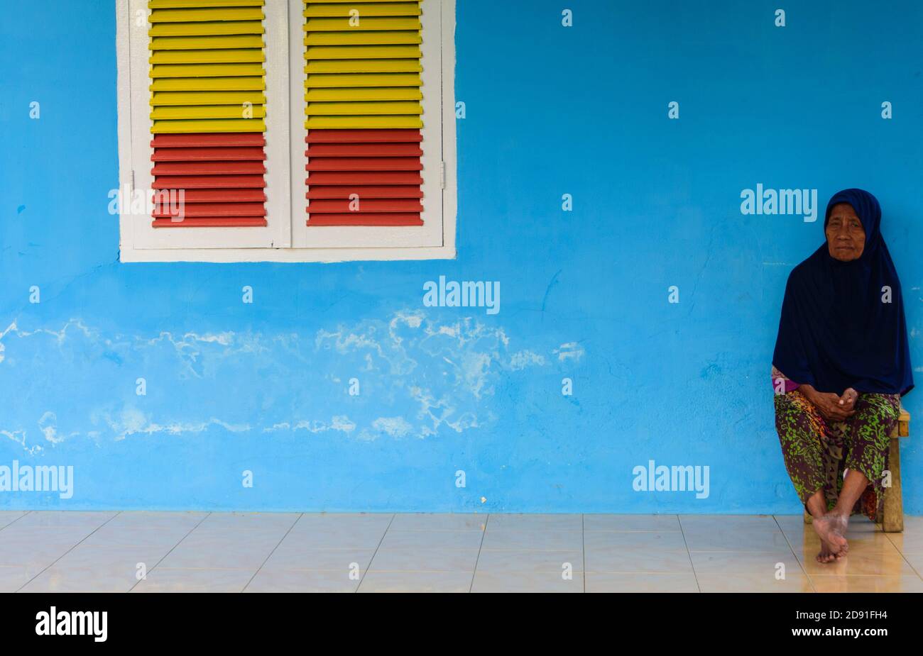 Donna seduta contro parete blu con persiane gialle/rosse, Ternate, North Molucche, Indonesia. Foto Stock