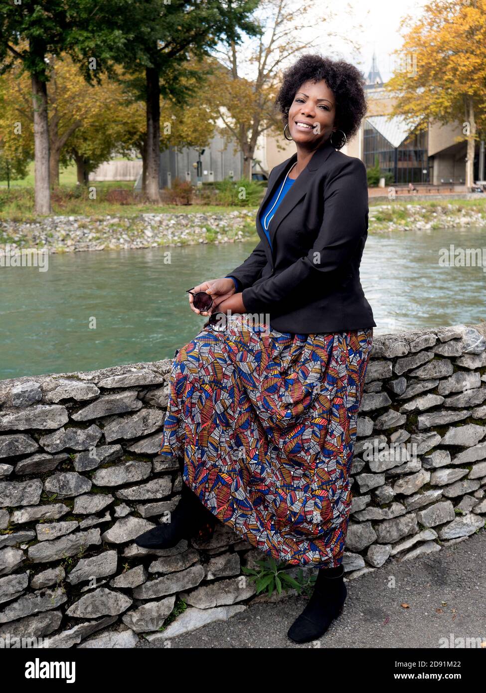 Ritratto verticale di una bella donna afroamericana sorridente che indossa una giacca nera e seduto vicino ad un fiume Foto Stock