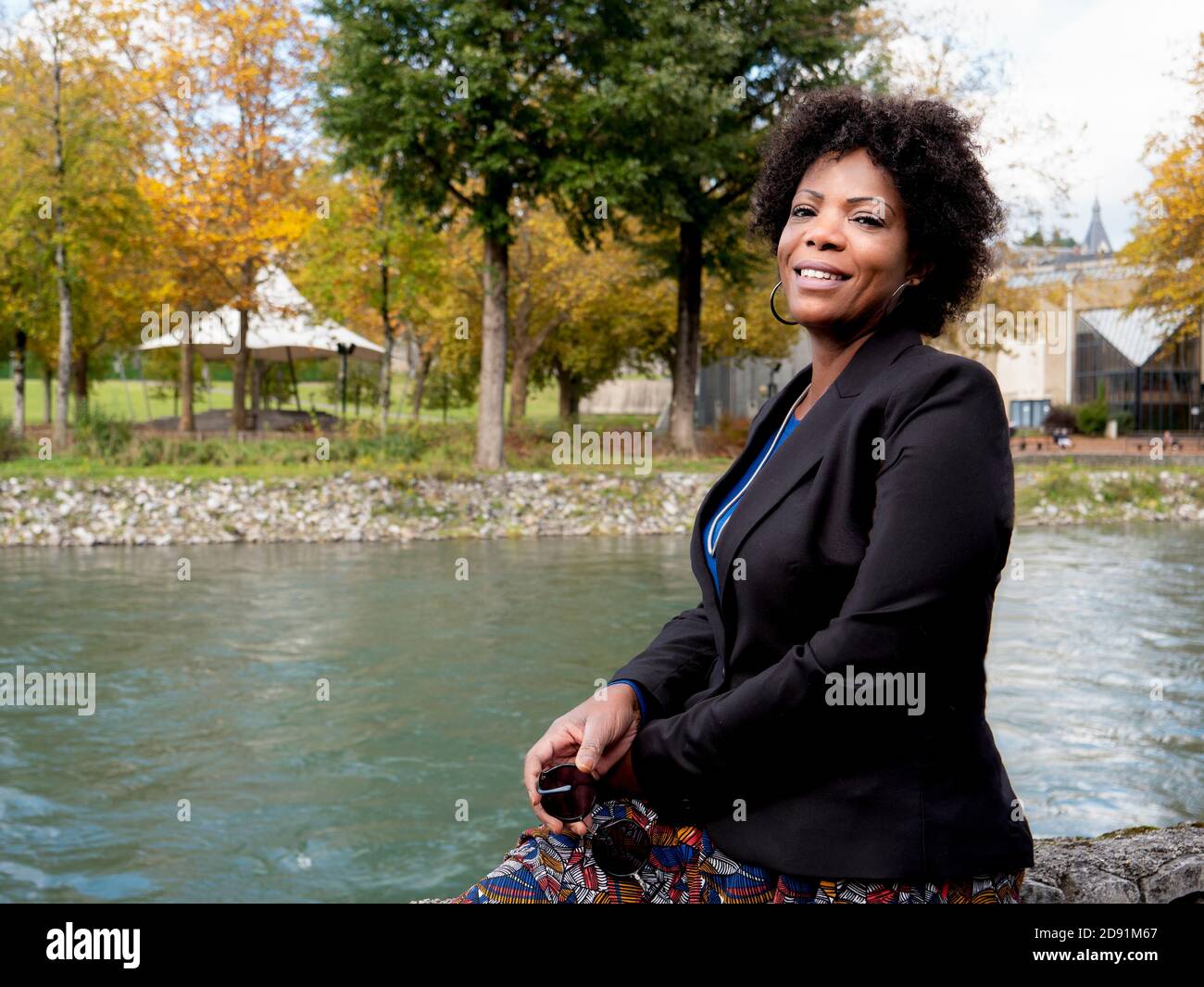 Ritratto orizzontale di una bella donna afroamericana sorridente che indossa una giacca nera e seduto vicino ad un fiume Foto Stock