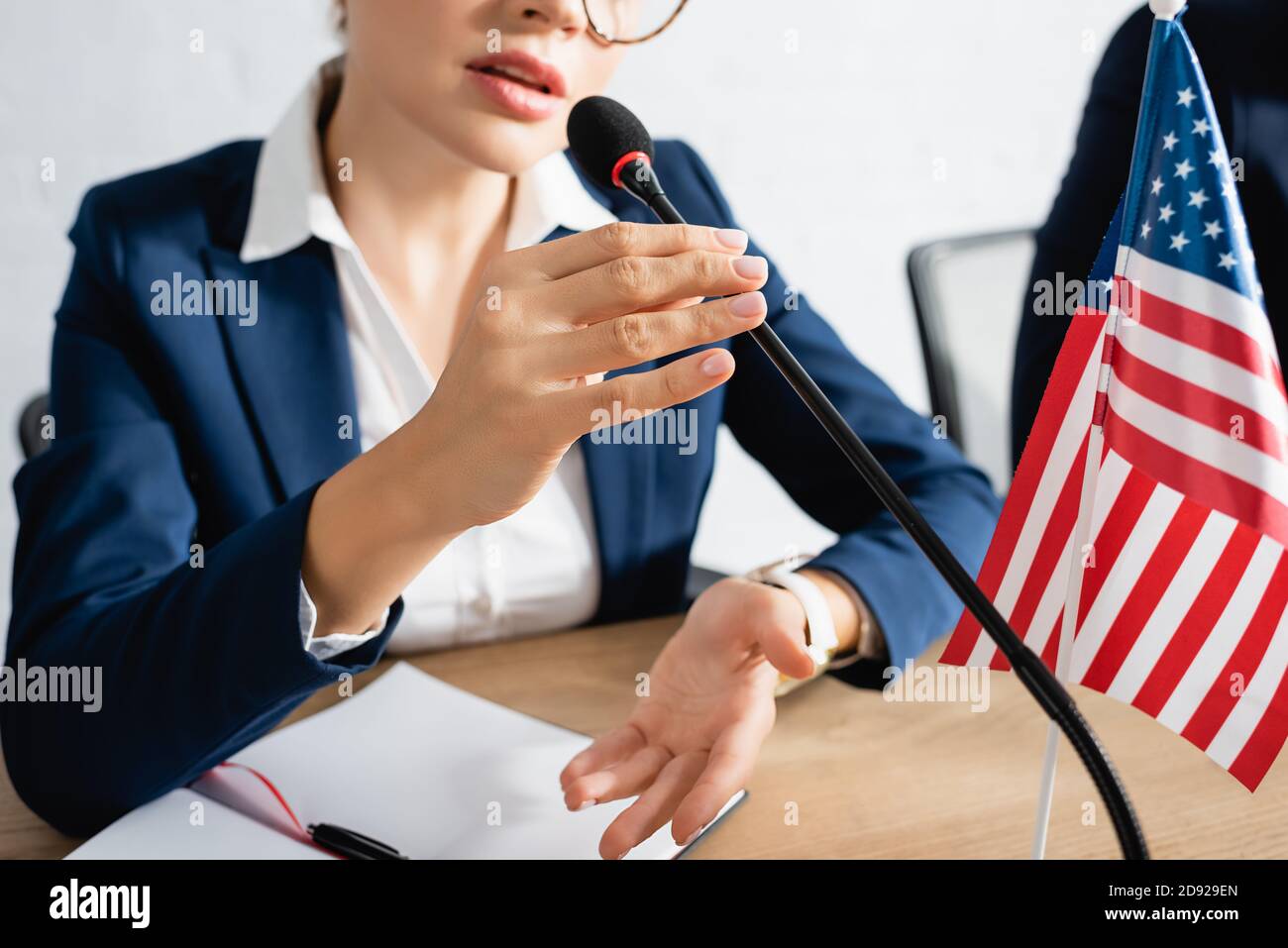Vista ritagliata della donna politico che parla in microfono, mentre si siede al tavolo vicino bandiera degli stati uniti in sala riunioni Foto Stock