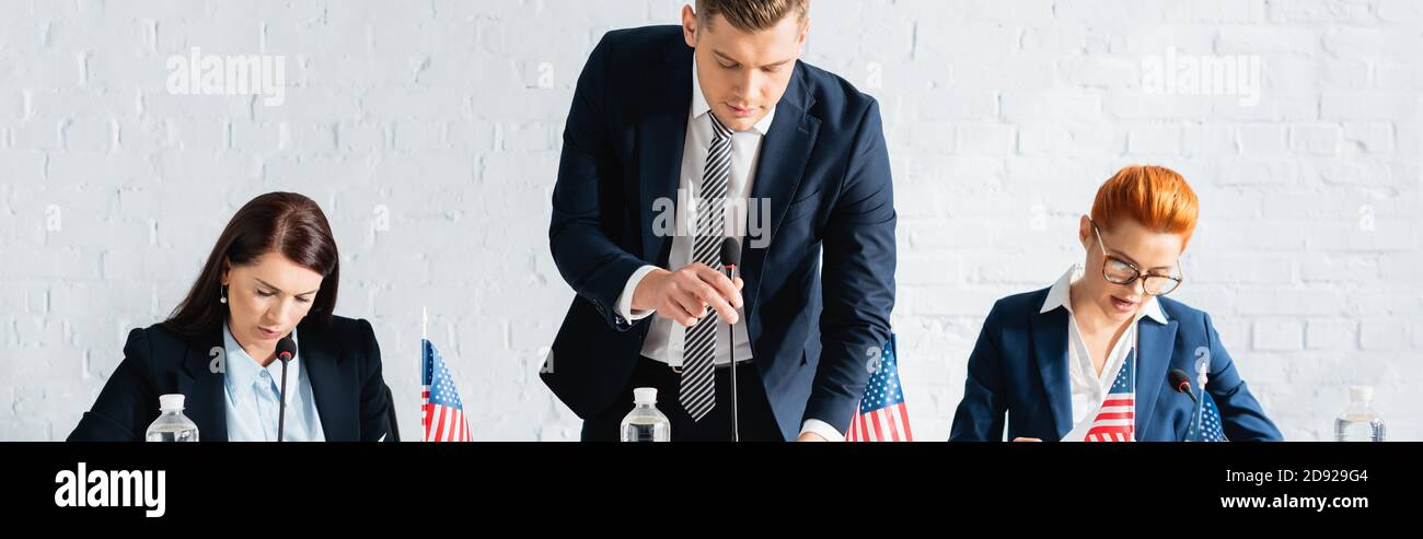 Uomo politico che si appoggia in avanti e tocca microfono vicino alle donne colleghi che lavorano in sala riunioni, banner Foto Stock