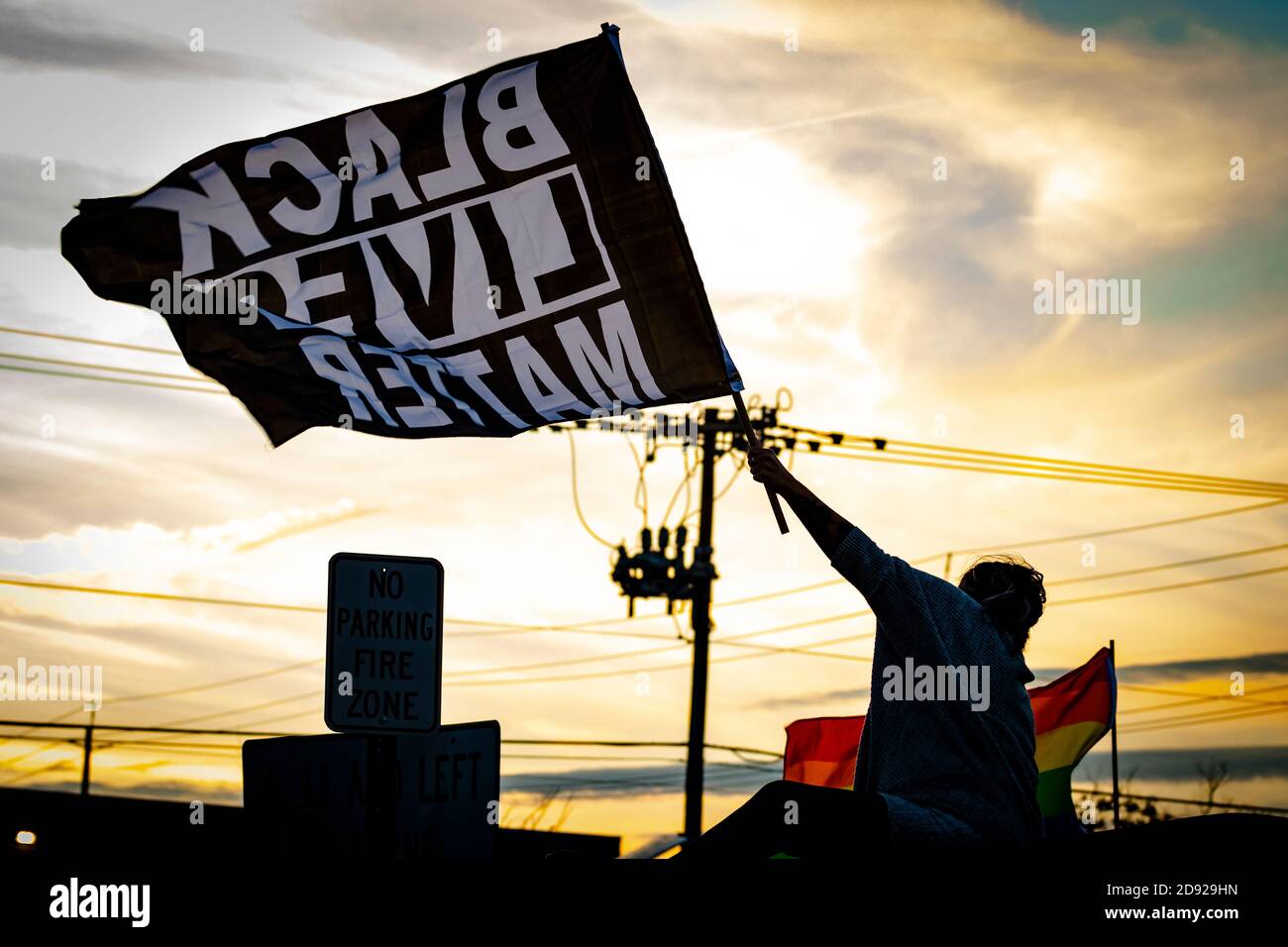 Silhouette scura al tramonto, di persona che tiene grande bandiera 'BLACK LIVES MATTER' sopra la testa al locale marciapiede rally; bandiera di orgoglio LGBT visto in lontananza. Foto Stock