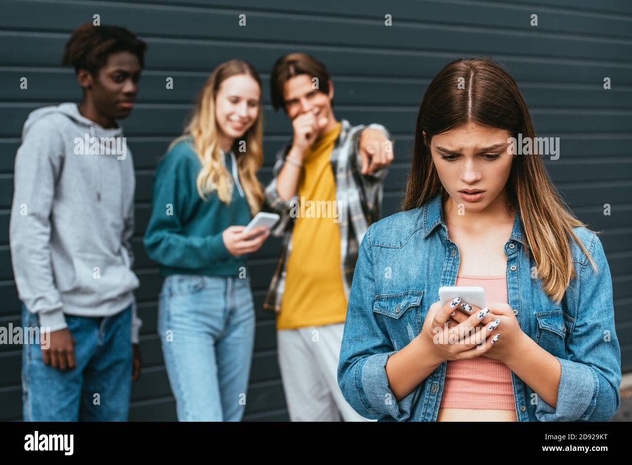 Fuoco selettivo di adolescente triste tenere smartphone vicino ridendo multietnico gli adolescenti puntano con le dita all'aperto Foto Stock