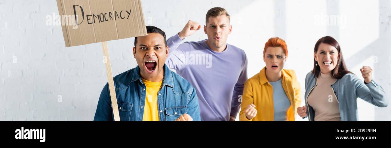 un uomo indiano aggressivo che tiene un cartello con scritta sulla democrazia vicino a urlare gente simile su sfondo sfocato, banner Foto Stock