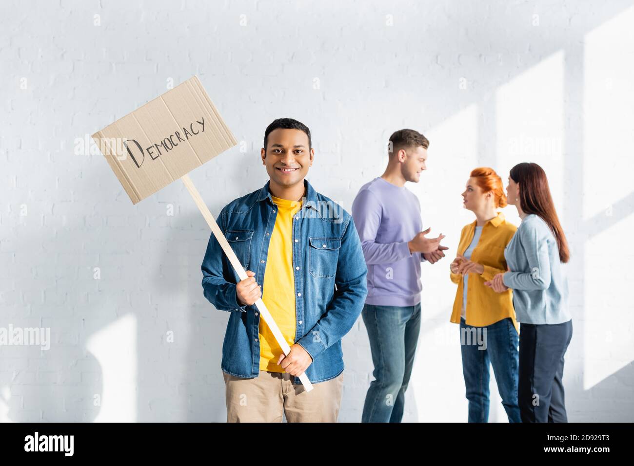 uomo indiano felice che tiene il cartello con la scritta democrazia mentre multiculturale persone con la stessa mentalità che parlano su sfondo sfocato Foto Stock