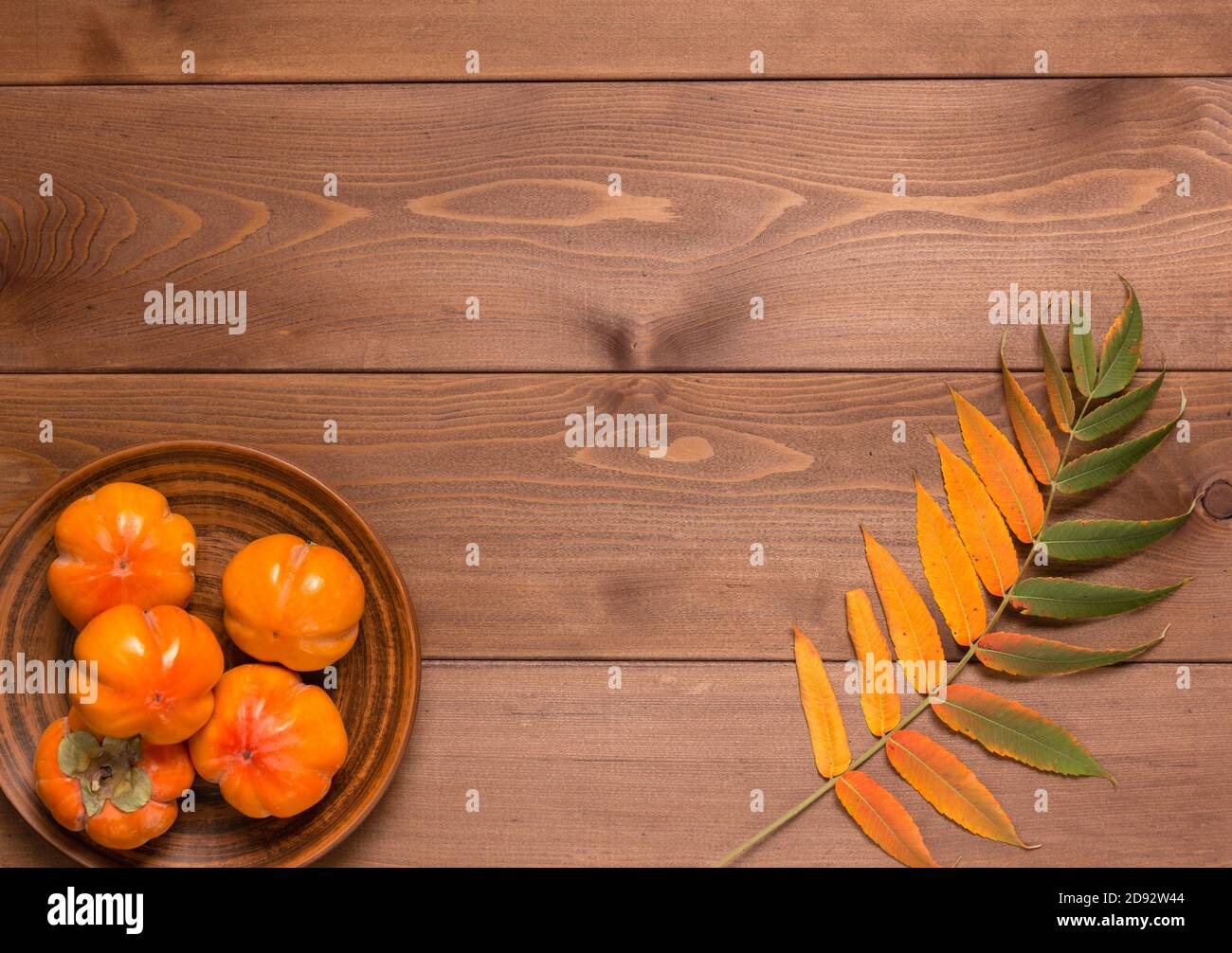 Abstract autunno ancora vita: Foglie giallite e persimmon maturo in una piastra di argilla su un tavolo di legno marrone. Disposizione piatta. Spazio di copia Foto Stock