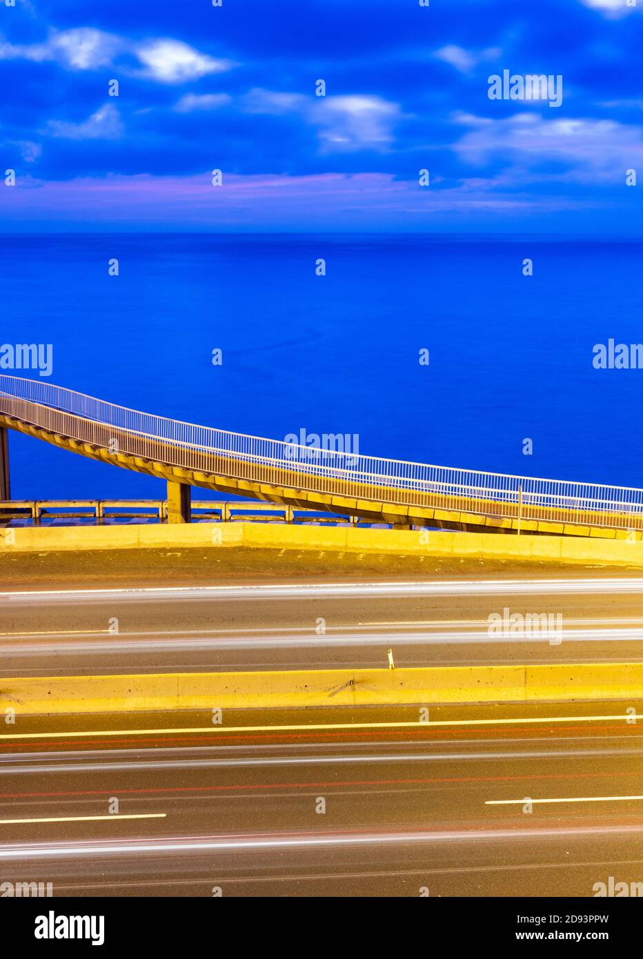 Lunga esposizione di sentieri di luce dal traffico sulla strada costiera con passerella e oceano in background all'alba. Foto Stock