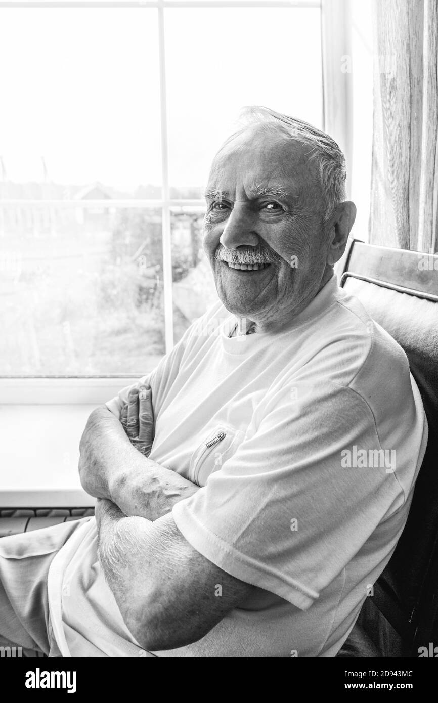 l'anziano è seduto su una sedia. Sorrido vecchio seduto su una sedia vicino alla finestra, ritratto, primo piano, immagine in bianco e nero Foto Stock