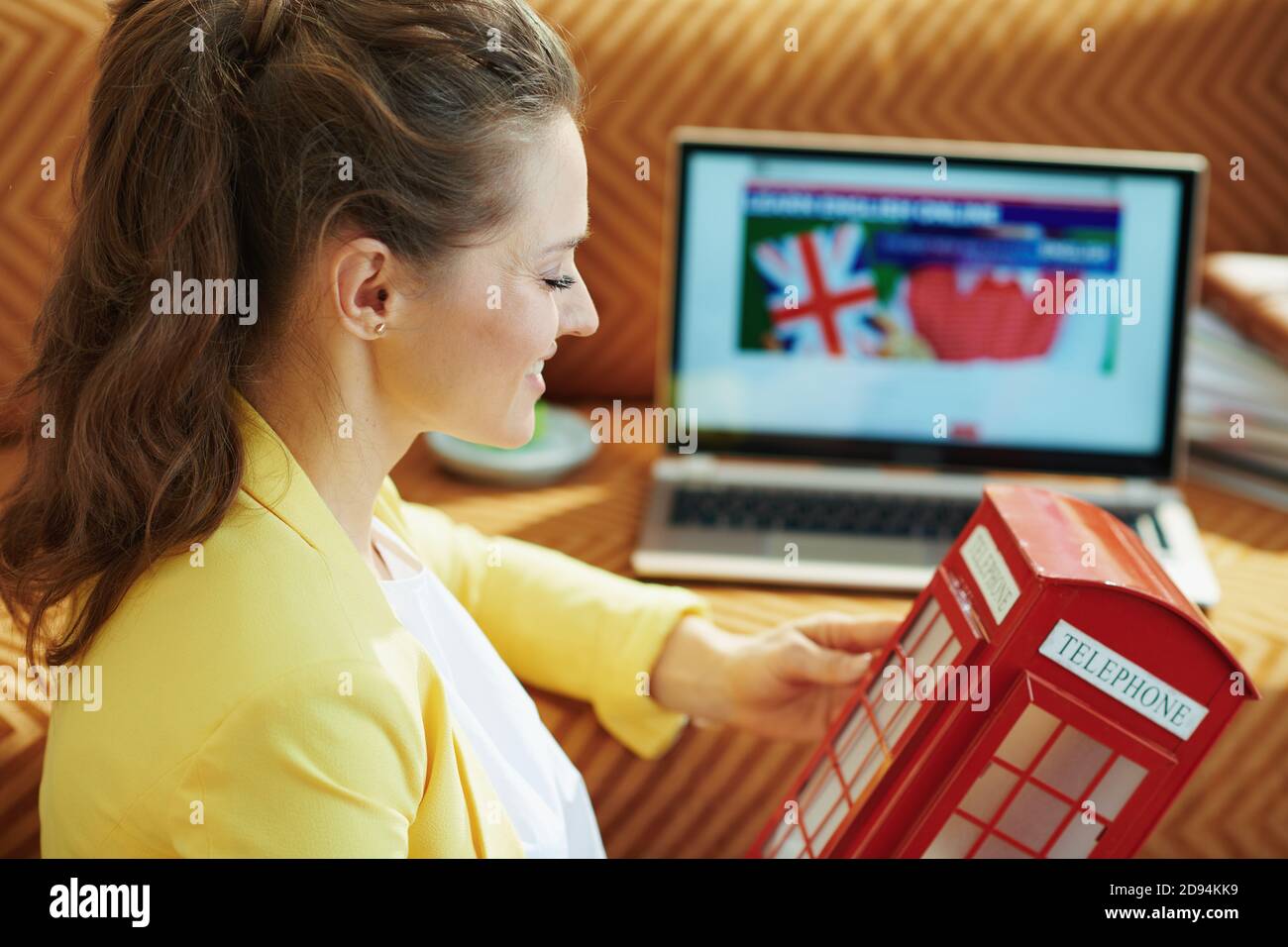 casalinga sorridente di 40 anni in giacca gialla con rosso cabina telefonica inglese imparare la lingua inglese online su un computer portatile nella casa moderna in s. Foto Stock