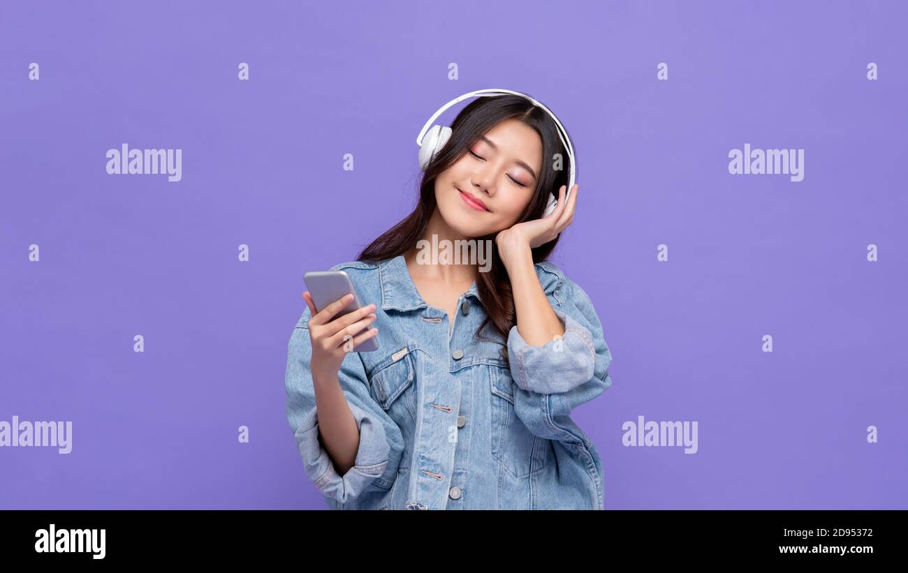 Ragazza asiatica felice indossando le cuffie ascoltare la musica dal cellulare telefono rilassante con gli occhi chiusi su sfondo viola isolato studio Foto Stock