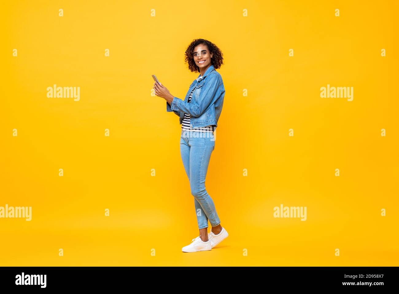 Ritratto a lunghezza intera di giovane donna afroamericana sorridente che guarda con la fotocamera mentre si tiene il telefono cellulare in uno studio isolato sfondo giallo Foto Stock