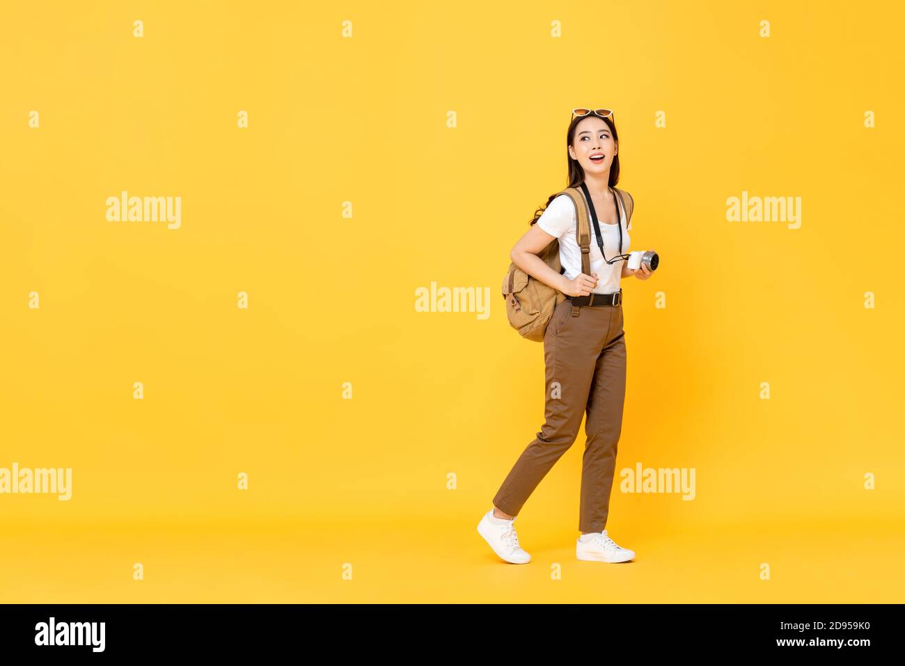 Ritratto a lunghezza intera di sorridente carino giovane turista asiatico con la telecamera che guarda lo spazio vuoto da parte su giallo isolato sfondo studio Foto Stock