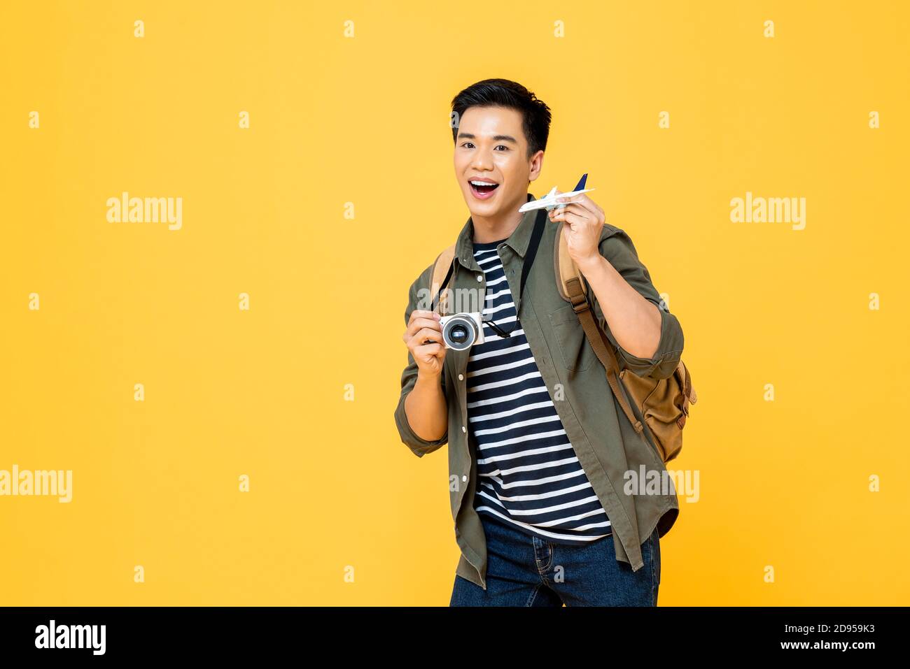 Ritratto di giovane bel sorriso asiatico maschio turista zaino in spalla modello di piano e telecamera isolati su sfondo giallo dello studio Foto Stock