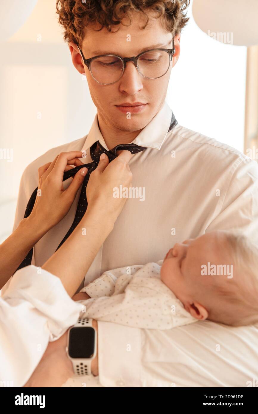 Donna che fissa i mariti legano mentre sta tenendo il loro figlio del bambino, in piedi a casa, ottenendo pronto per il lavoro Foto Stock