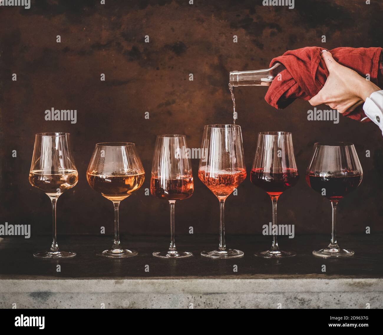 Sfumature di vino Rosa in calici con gambo messi in linea e womans mano versando il vino da bottiglia a bicchiere, arrugginito fondo marrone dietro. Enoteca, enoteca, degustazione Foto Stock