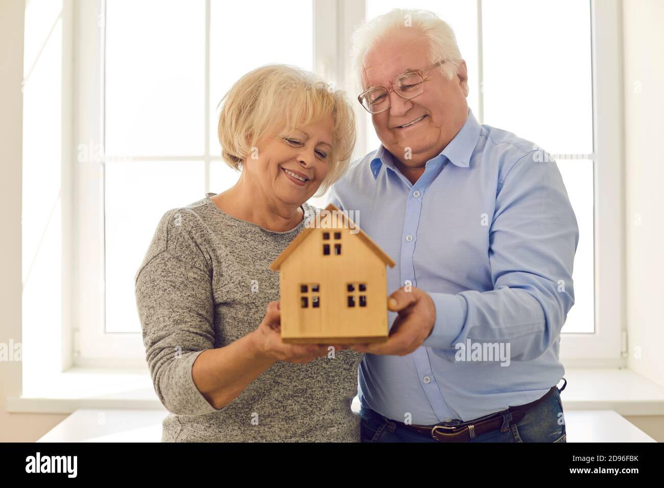 Sorridente coppia senior che tiene casa in miniatura in piedi vicino alla finestra nella loro nuova casa Foto Stock