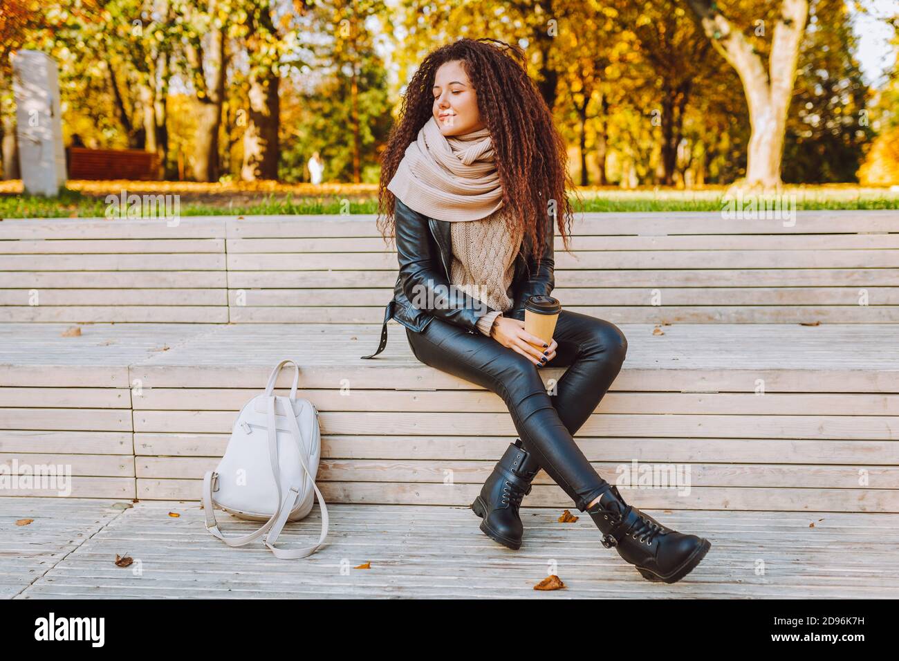 Donna afro positiva che indossa maglia maglione e sciarpa sedersi su panchina in autunno parco in giornata di sole con caffè e respirando in profondità Foto Stock