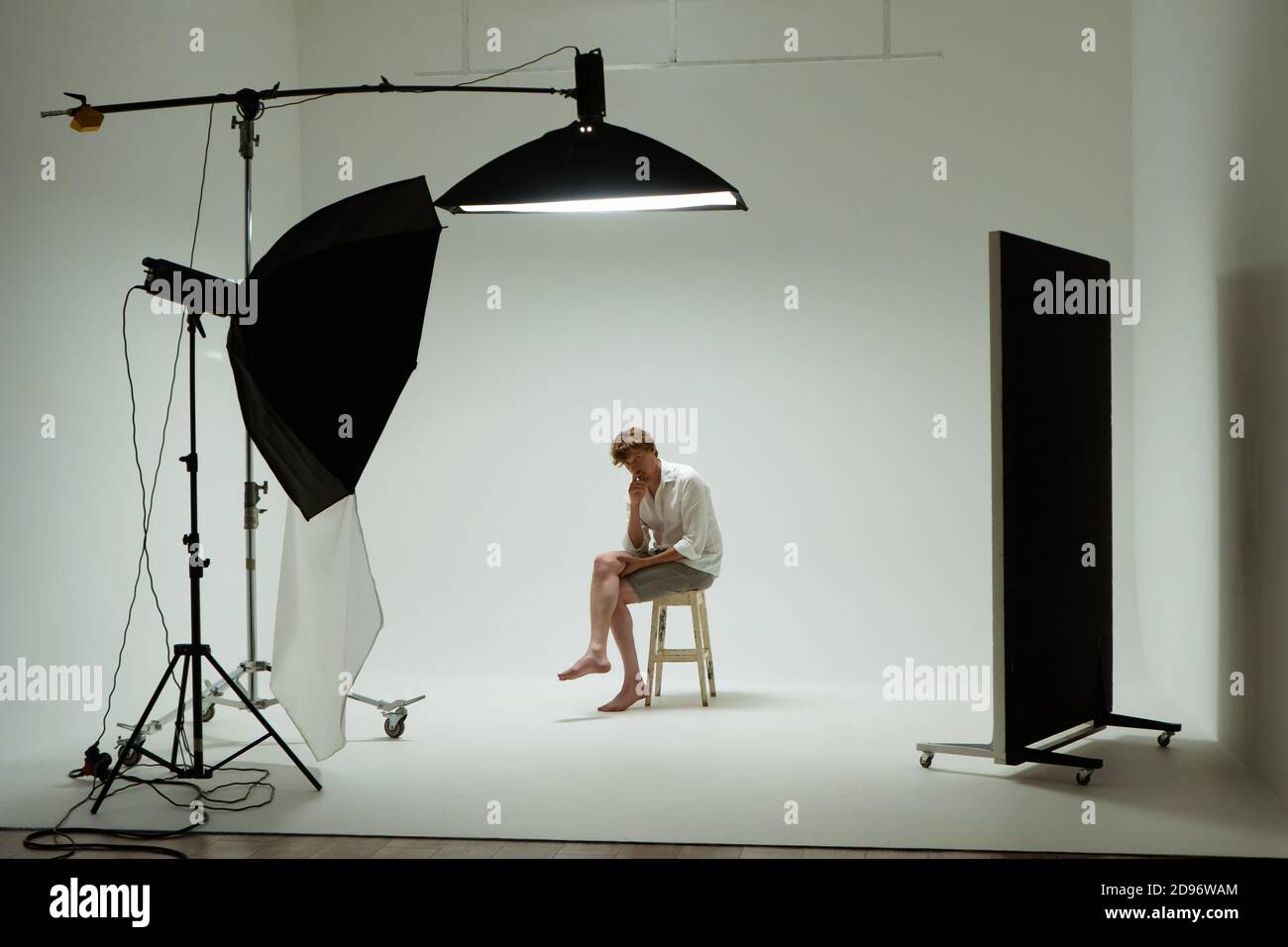 Il giovane uomo a piedi nudi si siede sulla sedia in posa di pensatore in posa in studio fotografico. Modello maschile con camicia bianca su sfondo bianco tra le attrezzature da studio Foto Stock