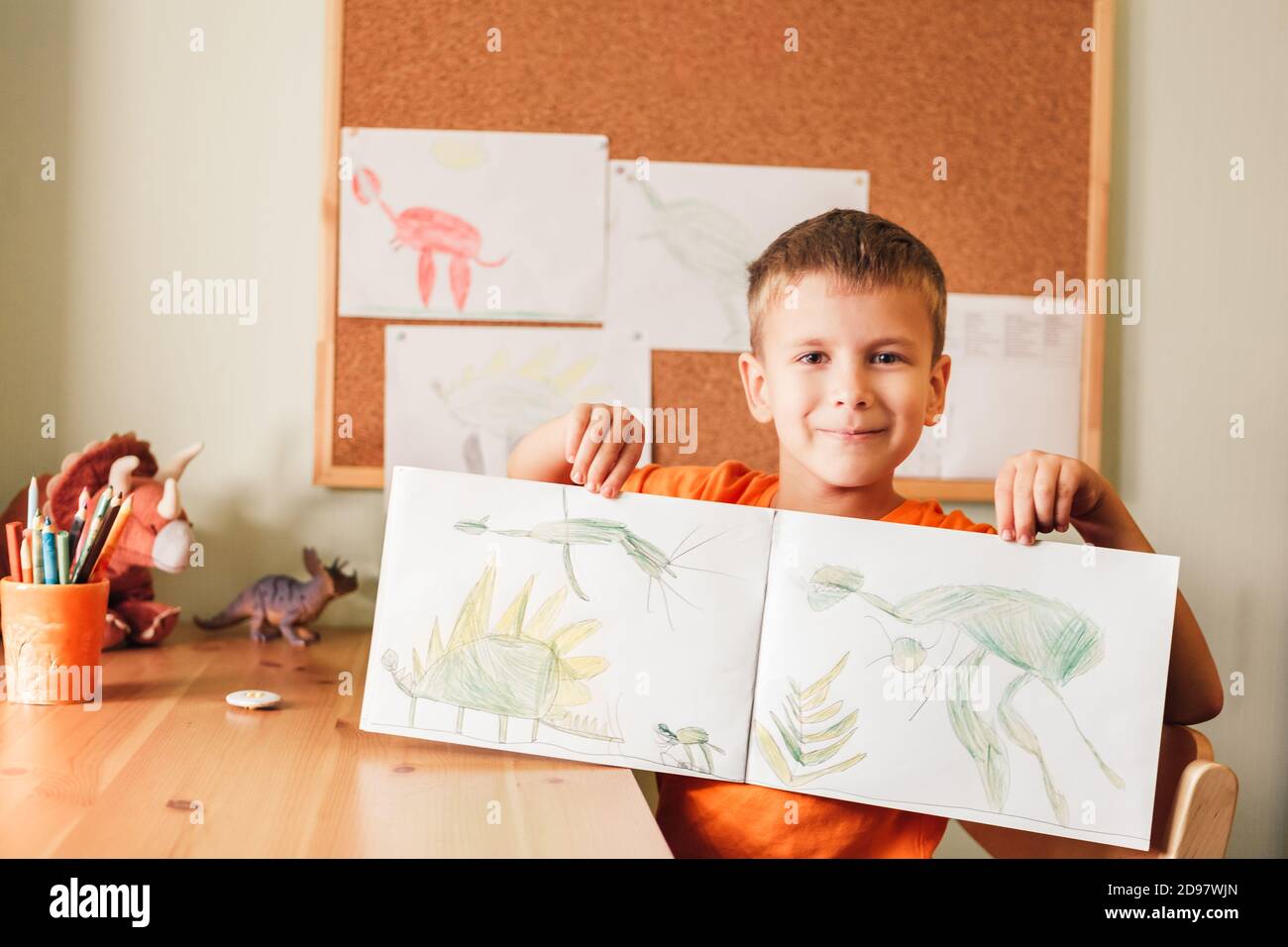 Cute bambino ragazzo disegno dinosauri su immagine con matite colorate seduto alla scrivania in camera Foto Stock