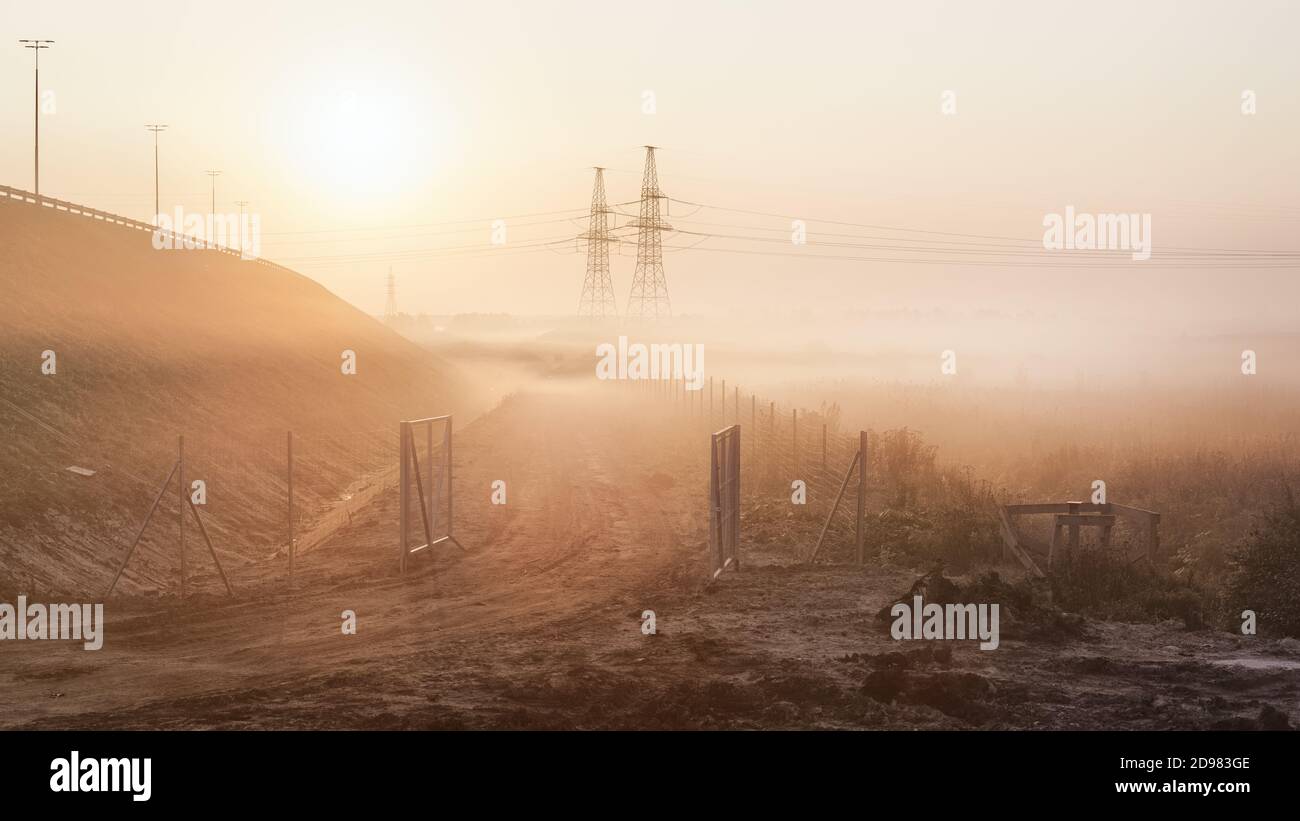 Paesaggio industriale all'alba foggy. Strada sterrata, autostrada e linea elettrica nella nebbia mattutina Foto Stock