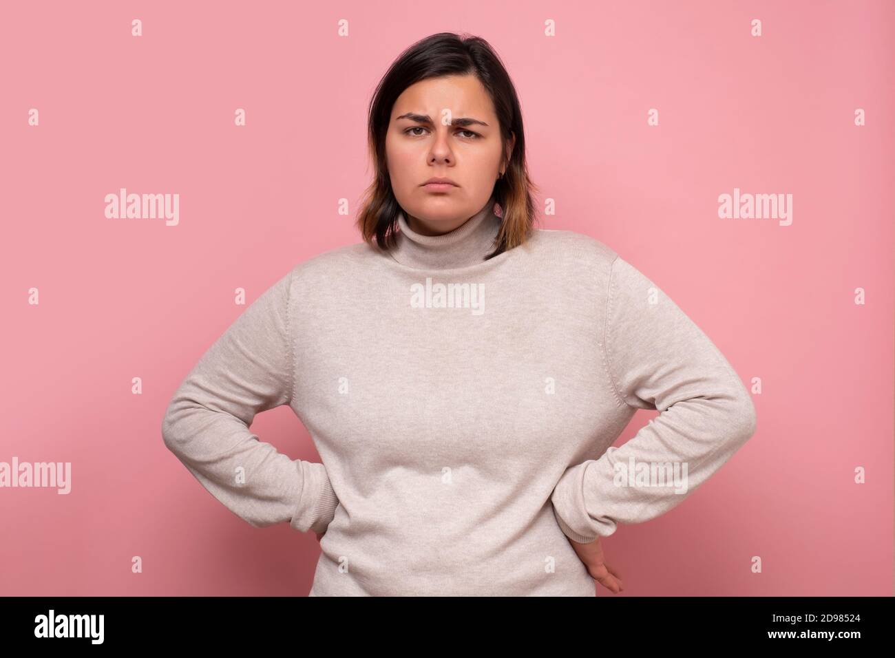 Donna seria osservante con le sue mani sui fianchi contro sfondo rosa Foto Stock