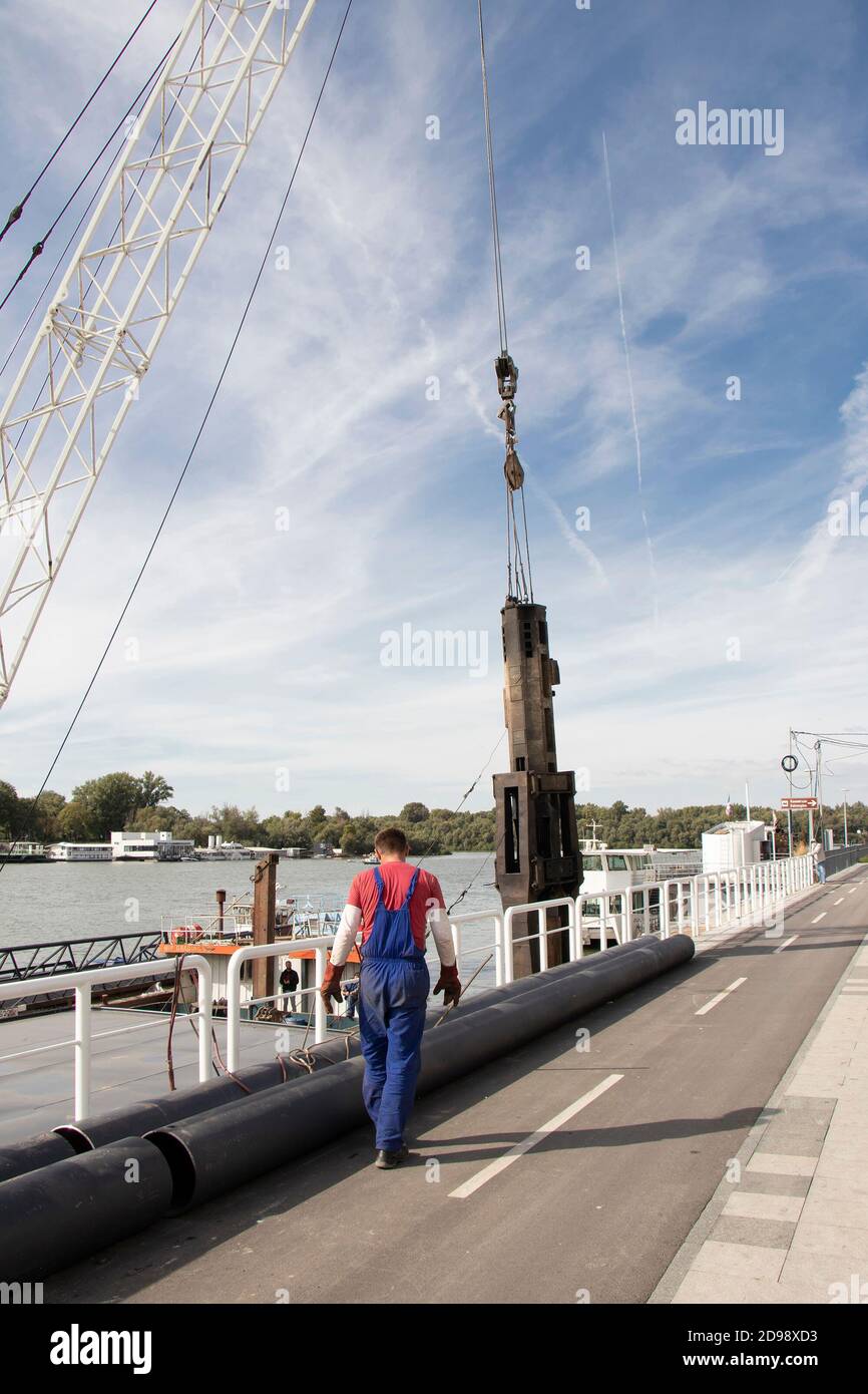 Belgrado, Serbia - 9 ottobre 2020: Gru di costruzione che solleva una grande punta industriale in acciaio per il pontile del fiume e un operaio, vista ad angolo basso riva del fiume Foto Stock