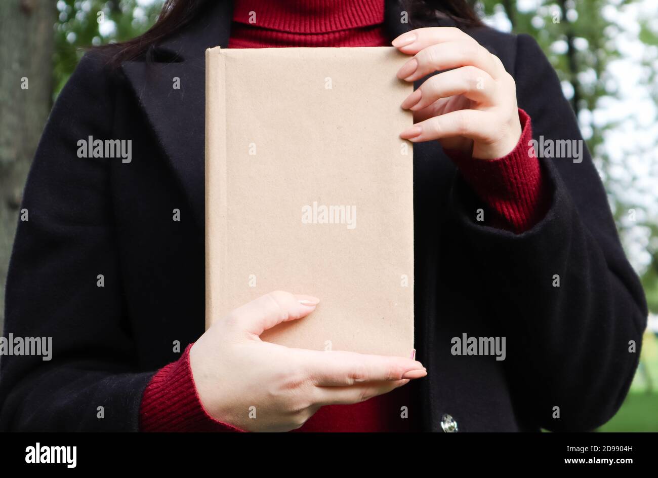 Vista ravvicinata della donna in camice di lana nera e maglione di borgogna che tiene un libro con copertina vuota nel parco. Spazio libero per il vostro concetto di lettura del libro Foto Stock