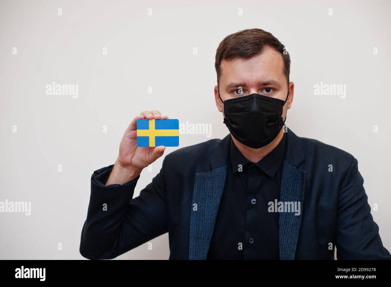 Uomo europeo indossare nero formale e proteggere maschera viso, tenere la carta bandiera svedese isolato su sfondo bianco. Europa coronavirus Covid concetto paese. Foto Stock