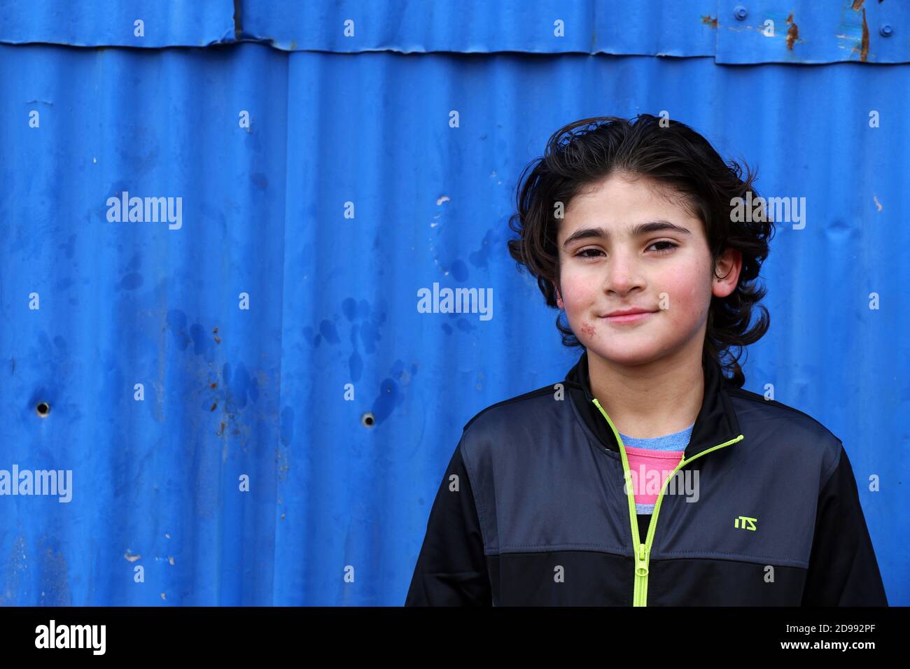 Un bambino sorride con ansia senza sapere cosa regge il futuro. Skaramagas ospita un campo profughi "temporaneo", una sorta di sala d'attesa dove i migranti provenienti principalmente da Siria, Iraq e Afghanistan soggiornano prima di essere reinsediati altrove in Europa. A Skaramagas, possono anche sapere se la loro richiesta di trasferimento è stata approvata o respinta. Makariou, Skaramagkas, Grecia. Foto Stock