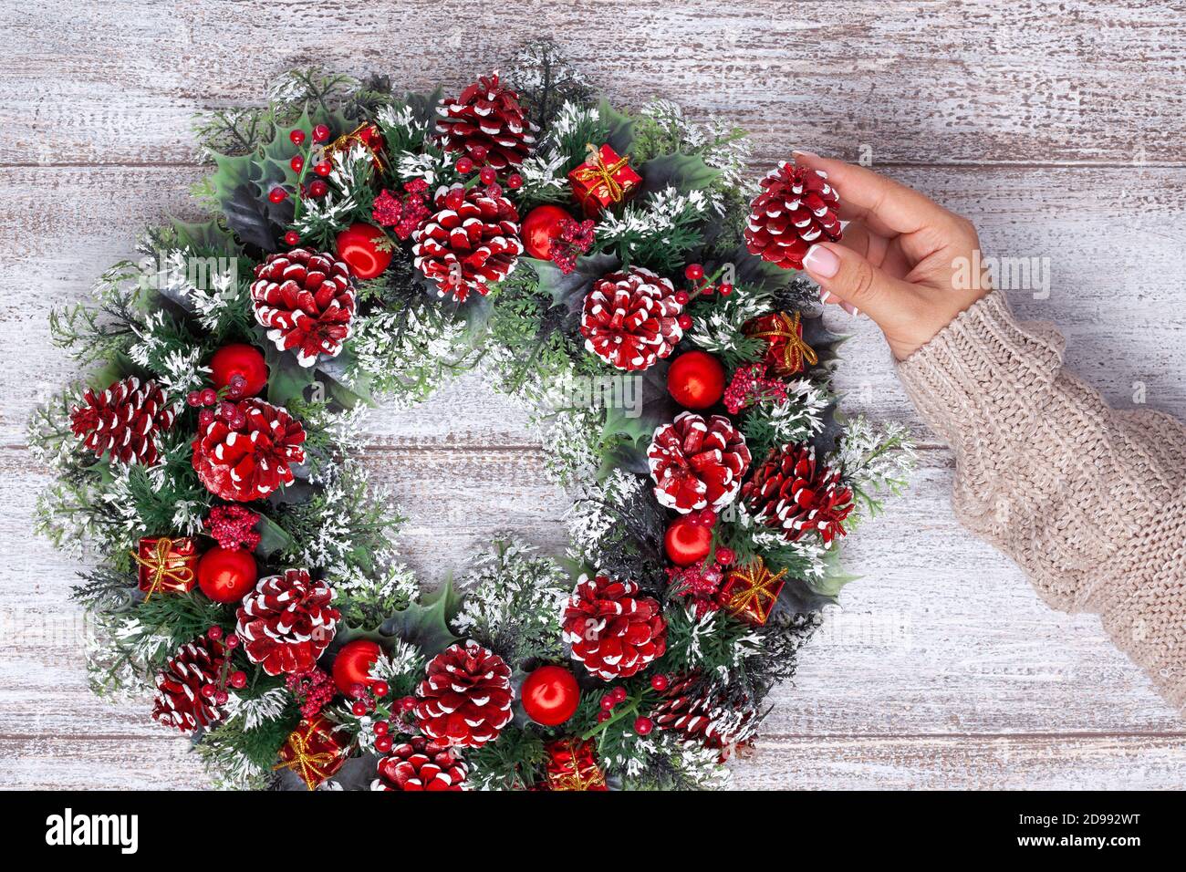 la mano femminile in un pullover beige lavorato a maglia aggiunge un rosso Ha verniciato il bump ad una corona di Natale fatta in casa su un legno sfondo Foto Stock