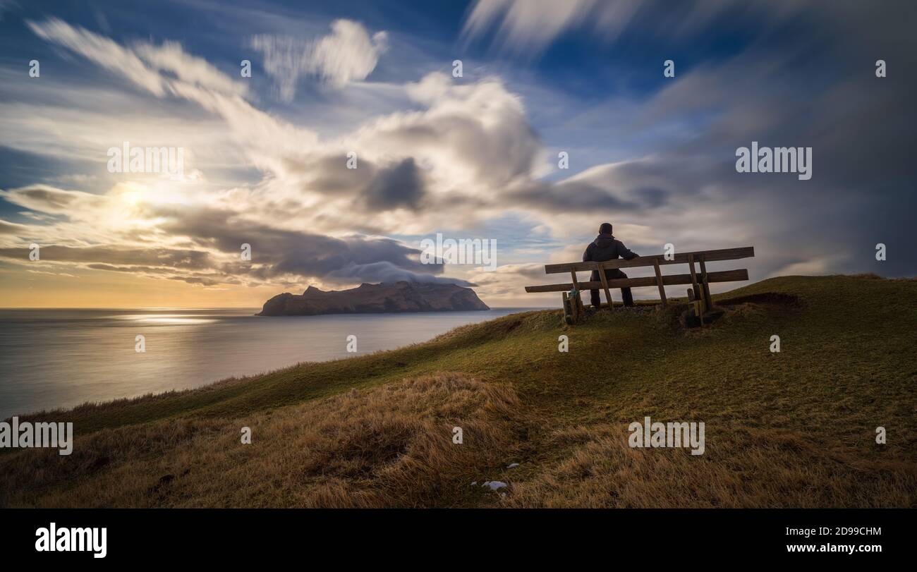 Uomo in panchina al villaggio di Gasadalur e isola di Mykines al tramonto, Isole Faroe Foto Stock