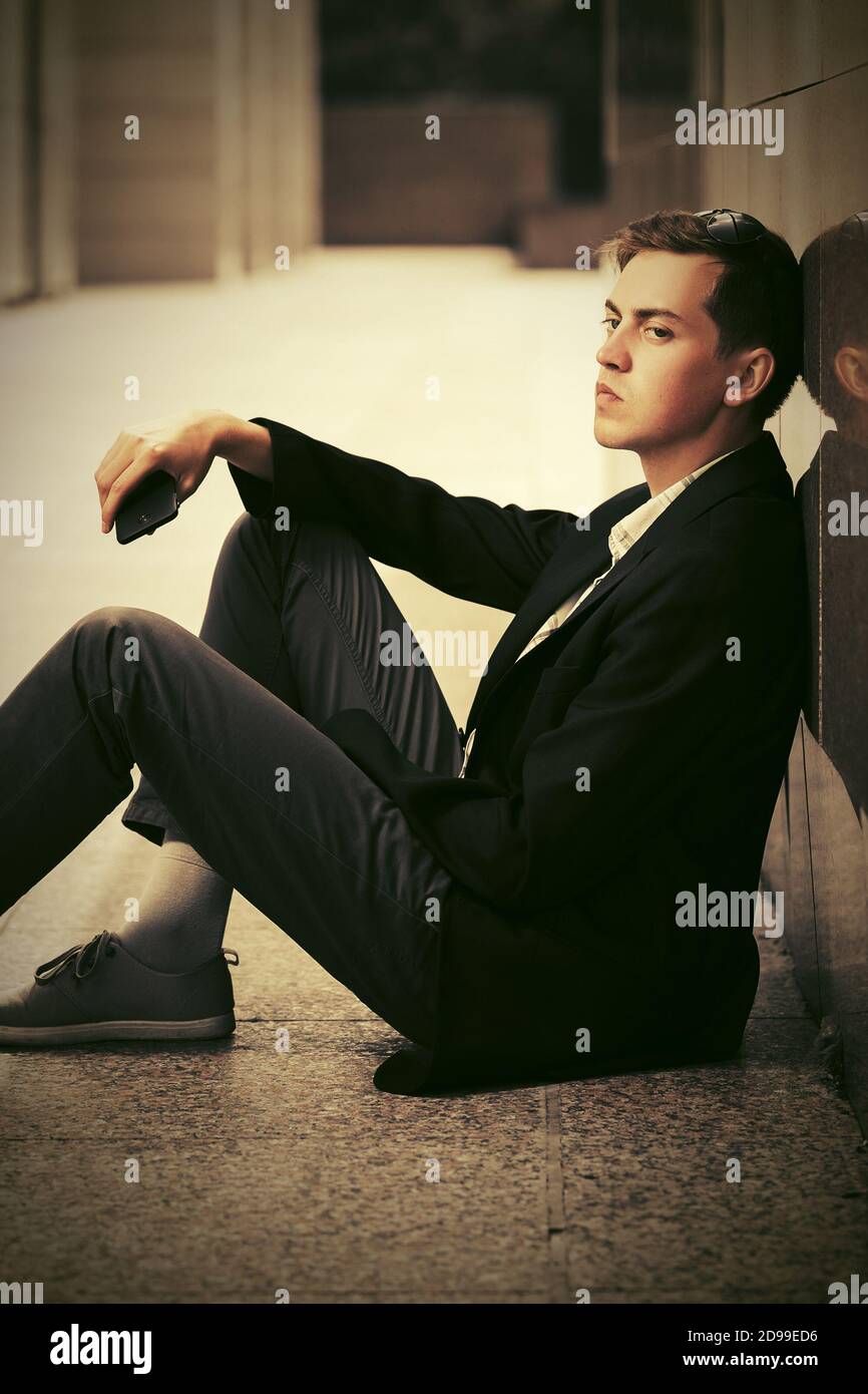 Giovane uomo con telefono cellulare seduto al muro street elegante modello maschile in blazer nero e pantaloni grigi Foto Stock